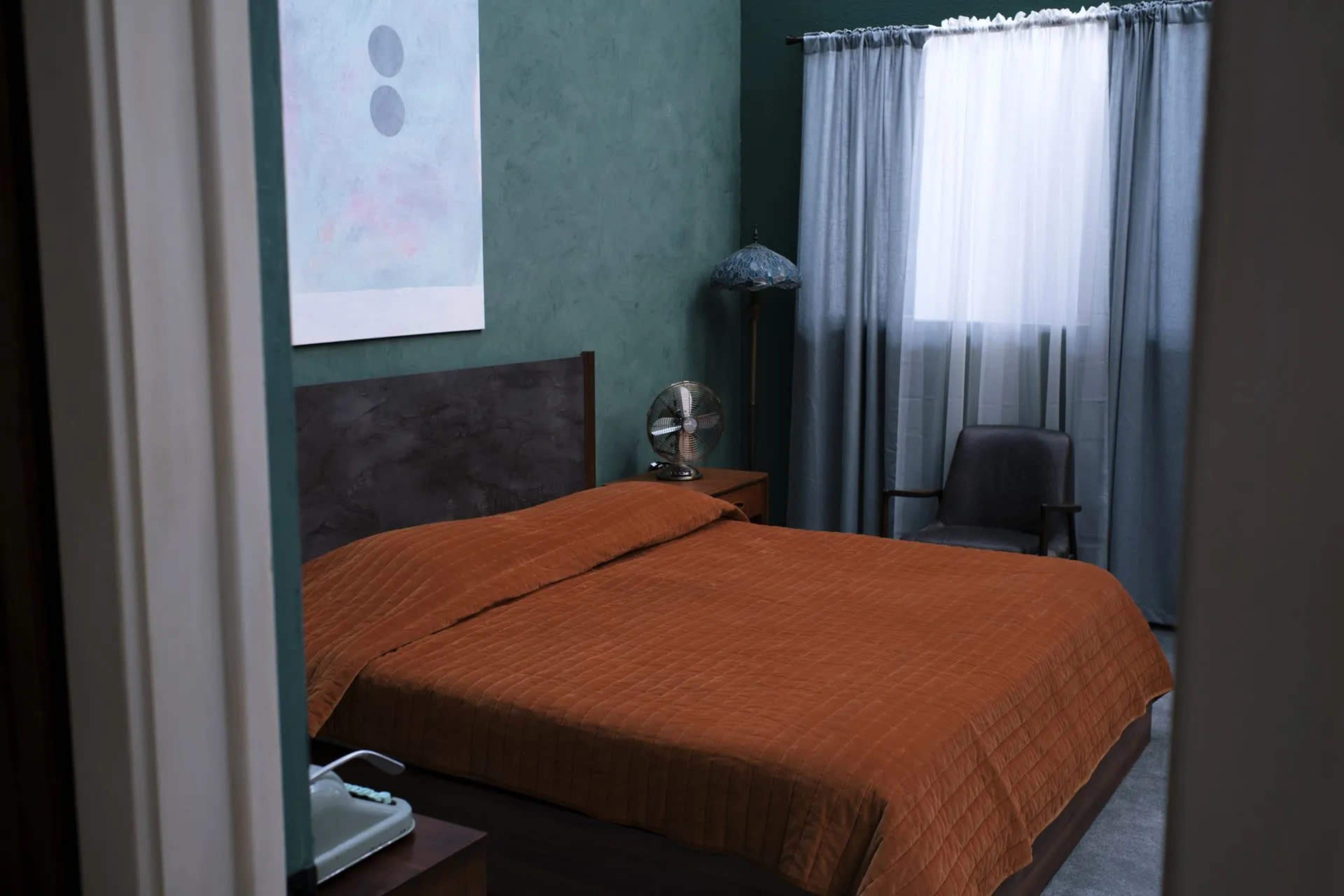 The image shows a bedroom with a large bed covered in an orange quilt, a window with sheer curtains, a bedside table with a lamp, and an armchair in the corner.