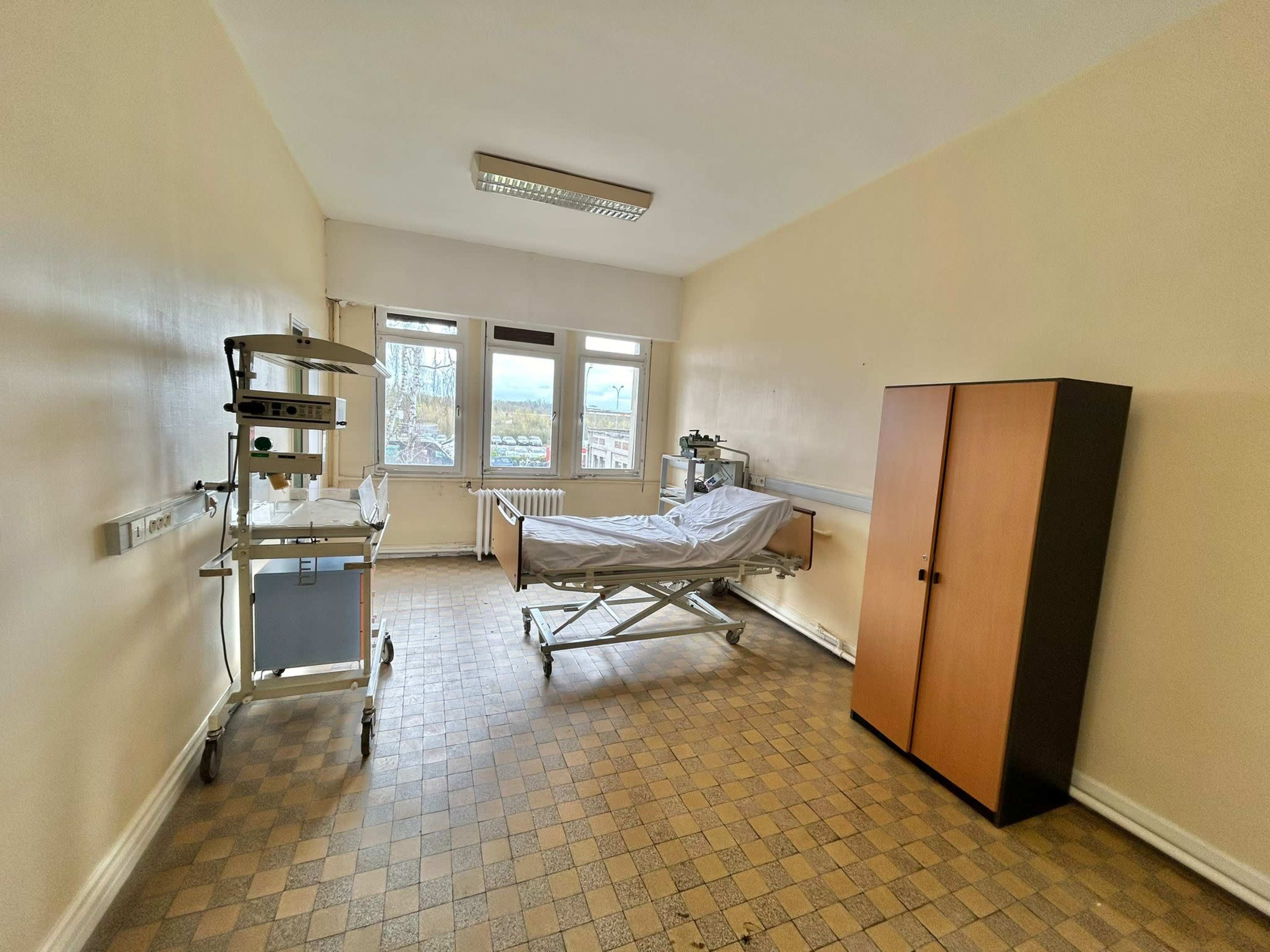 A hospital room with a bed, a bedside table, a medical cart, and a wardrobe against a wall with windows.