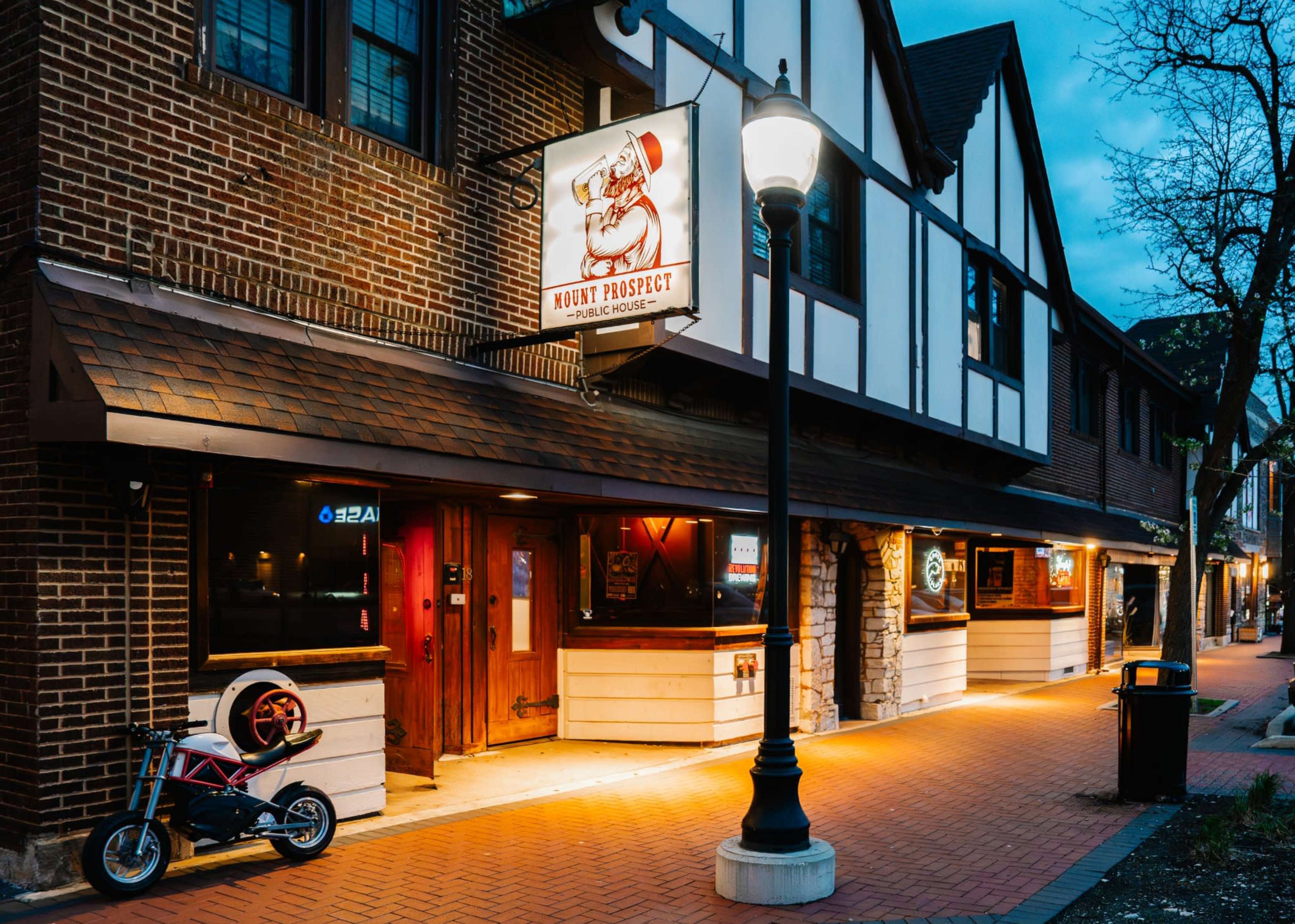 The image shows a brick building with a traditional facade, featuring a sign for "Muddy Prospect" and a small motorcycle parked outside, illuminated by a nearby streetlamp at dusk.