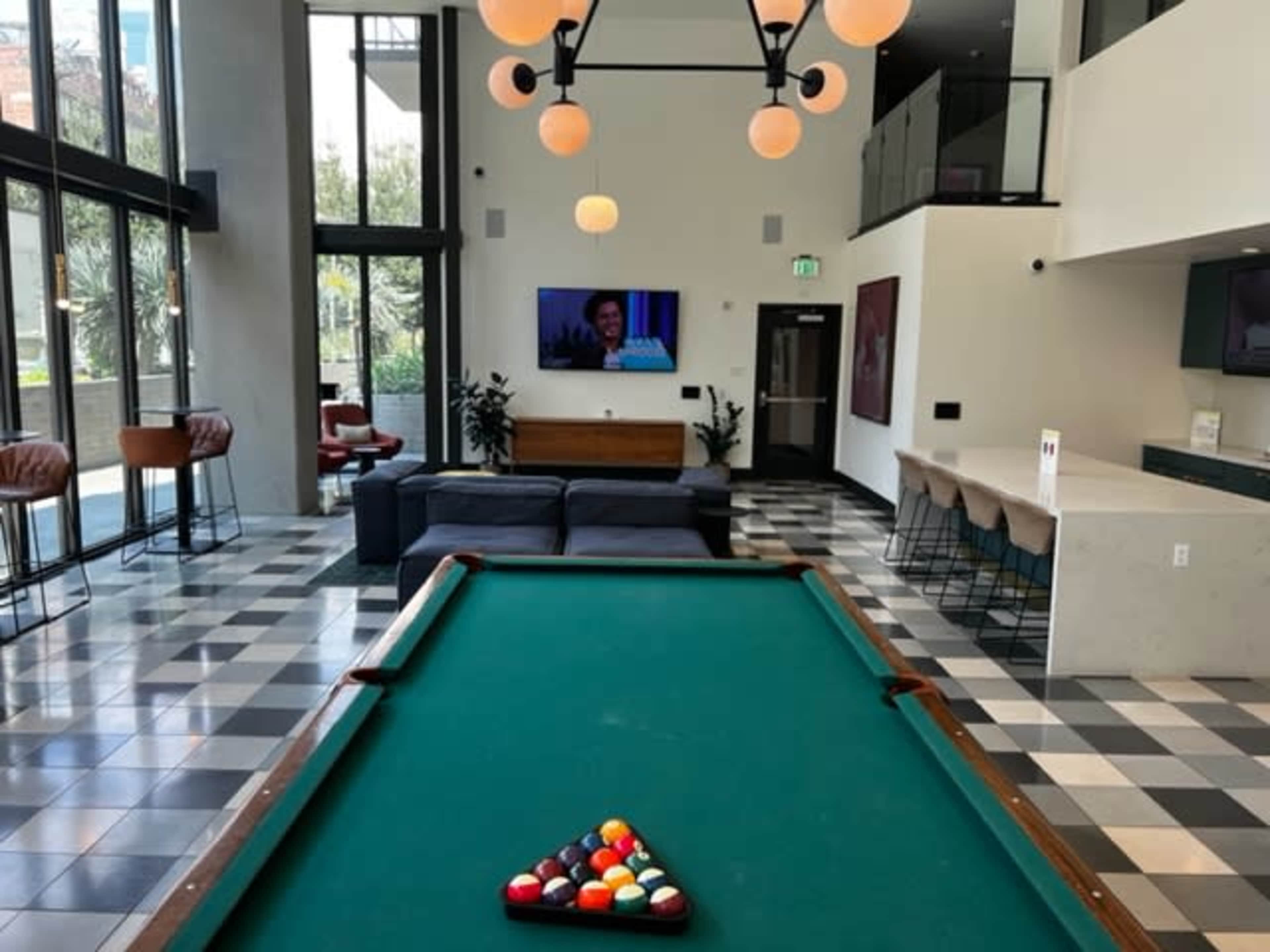 The image shows a recreational area featuring a pool table in the foreground, a seating area with couches, and a television mounted on the wall.