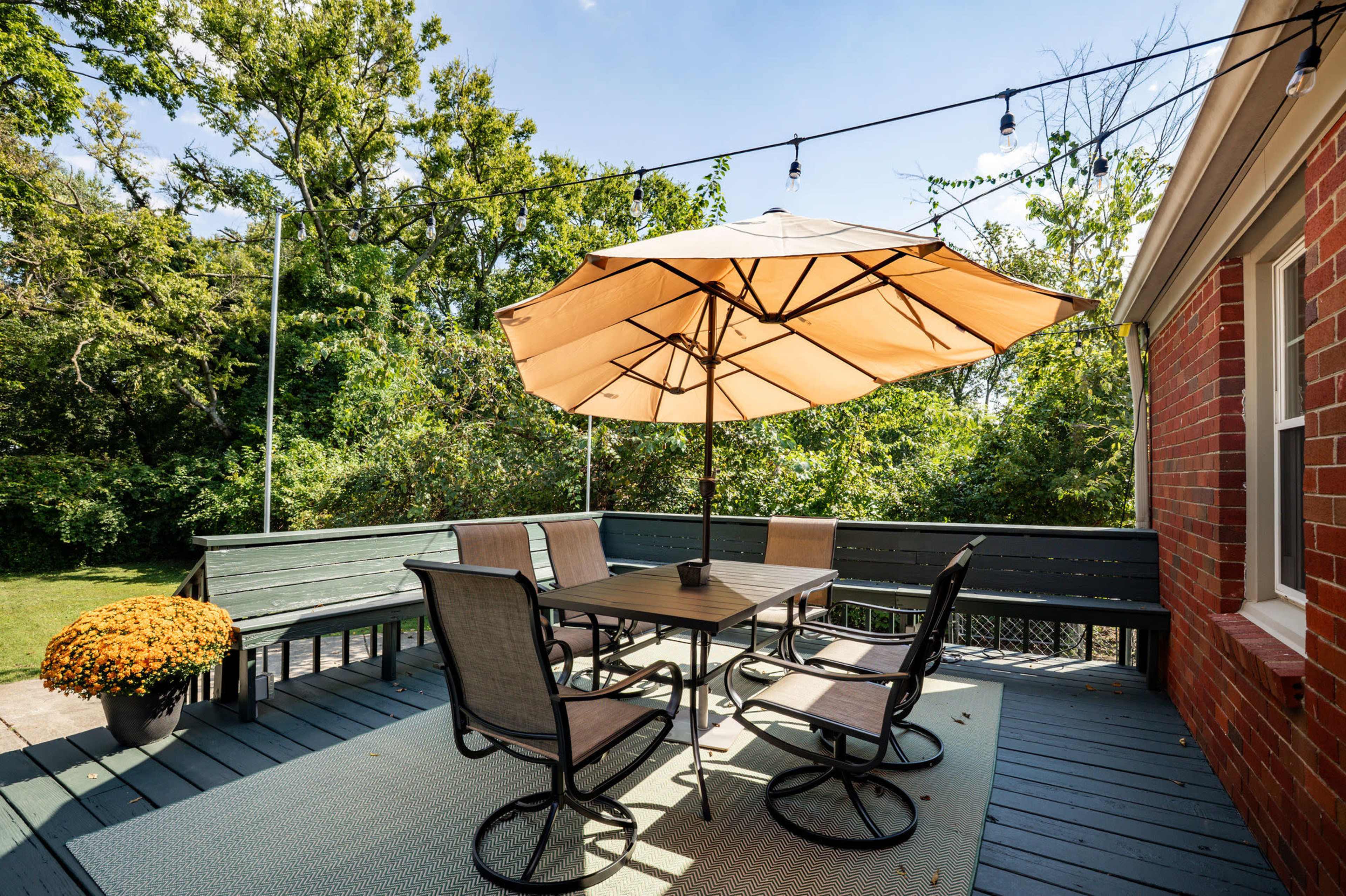 A patio features a table surrounded by six chairs, an umbrella providing shade, and a potted plant adorned with yellow flowers.