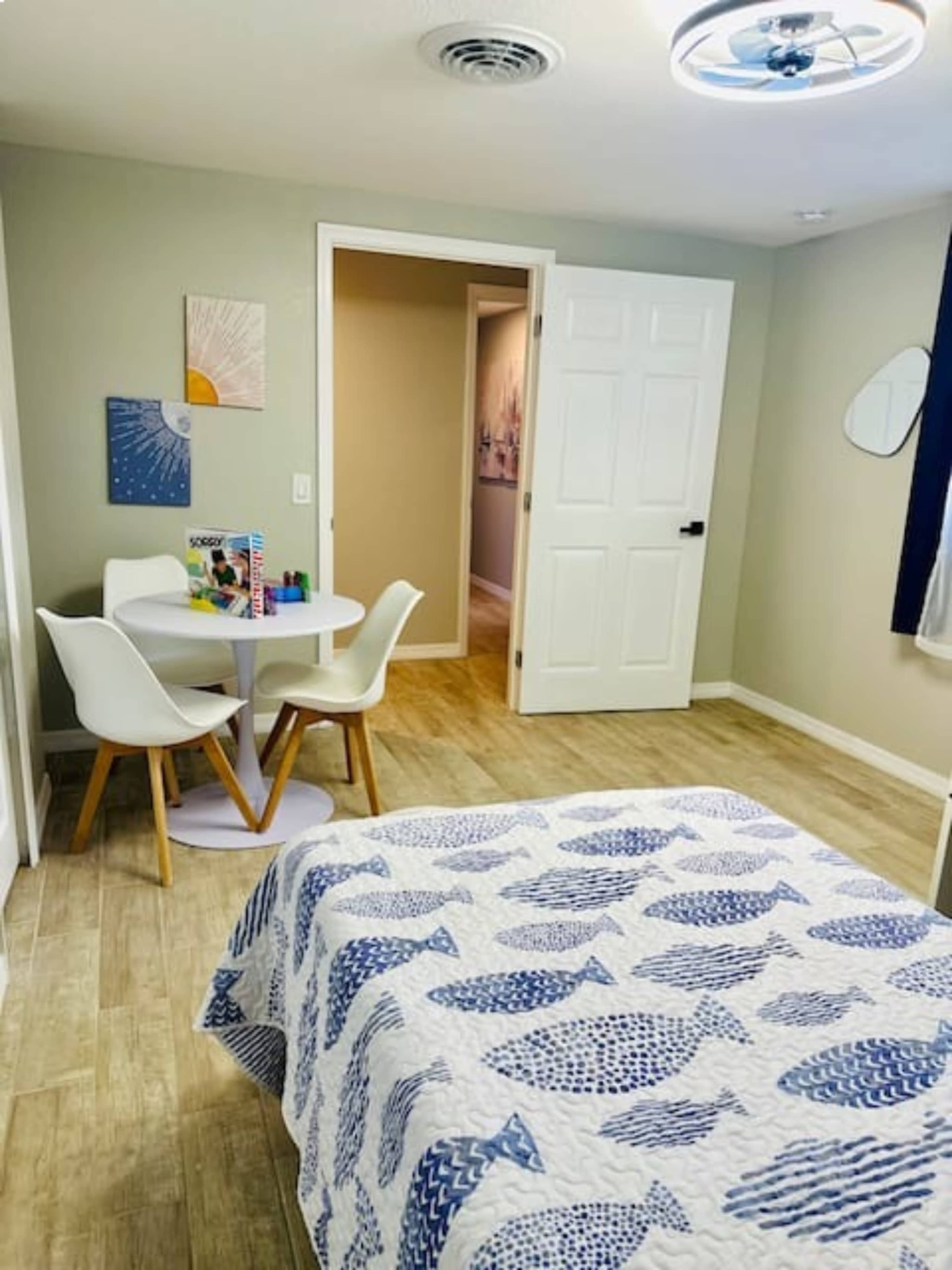 A bedroom with a blue and white patterned quilt on the bed, a small round table with chairs, and a doorway leading to another room.