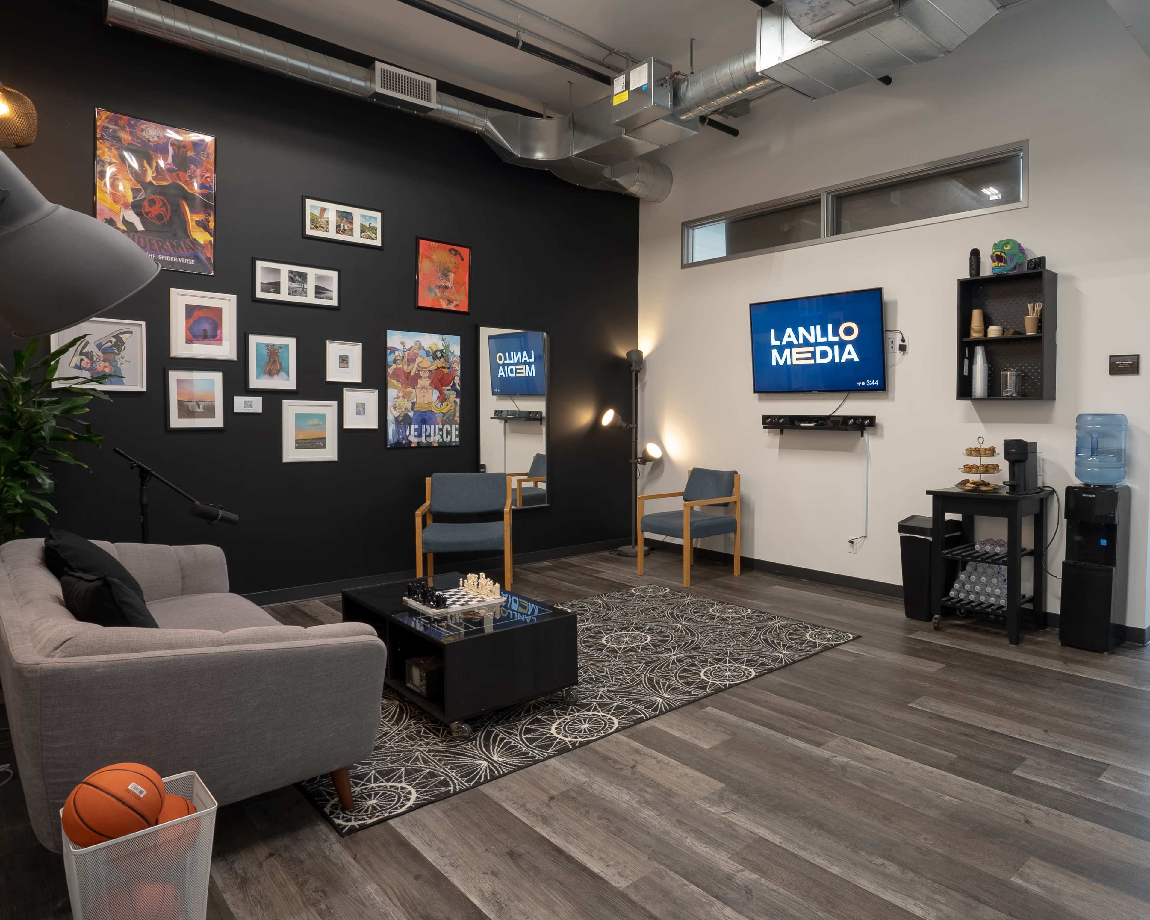 A modern lounge area features a grey sofa, two chairs, wall art, a television displaying "LANLLO MEDIA," and a water cooler, all set against a black accent wall.