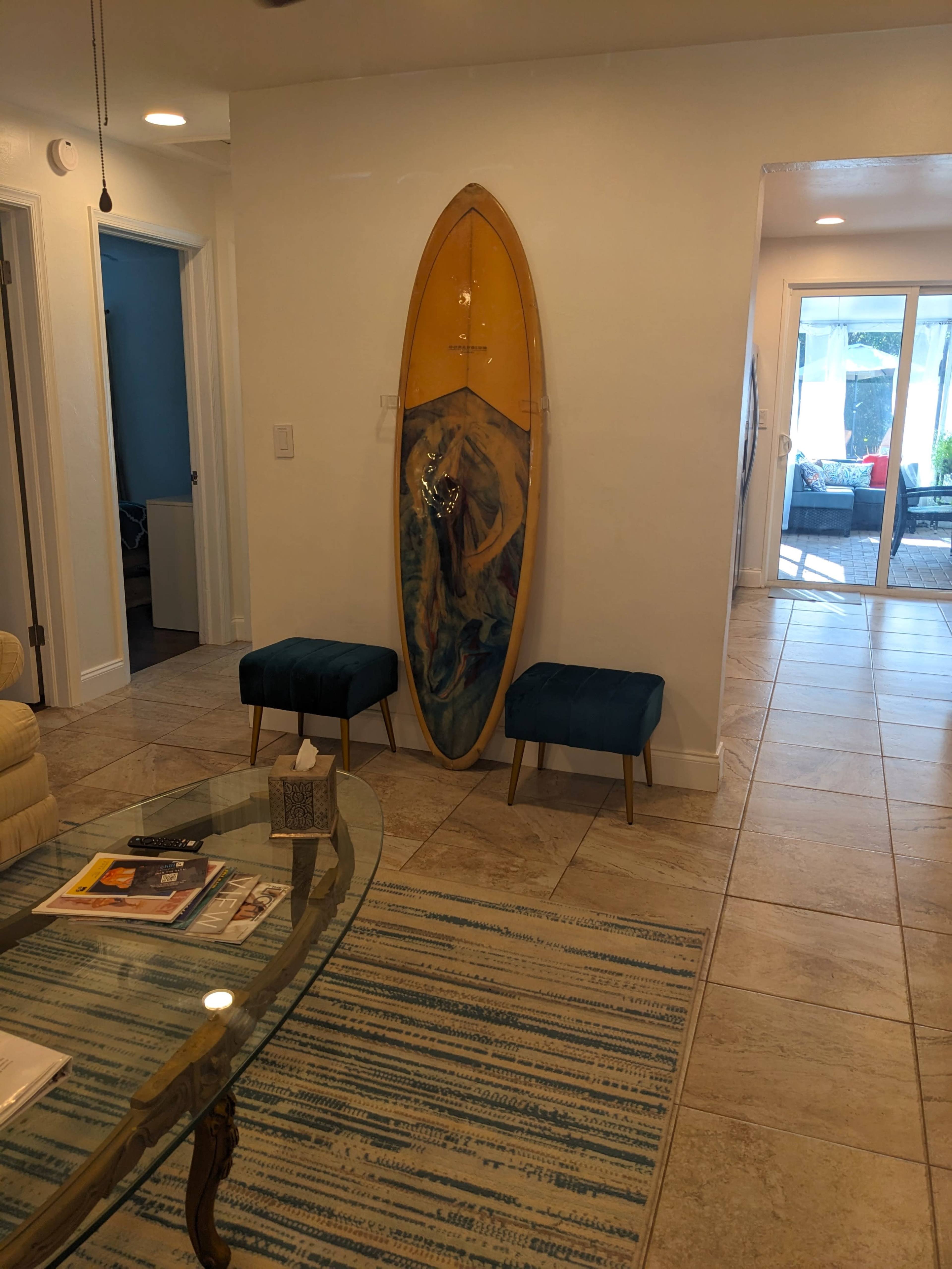 The scene features a surfboard mounted on the wall next to two teal ottomans, with a glass coffee table and patterned rug in a well-lit hallway.