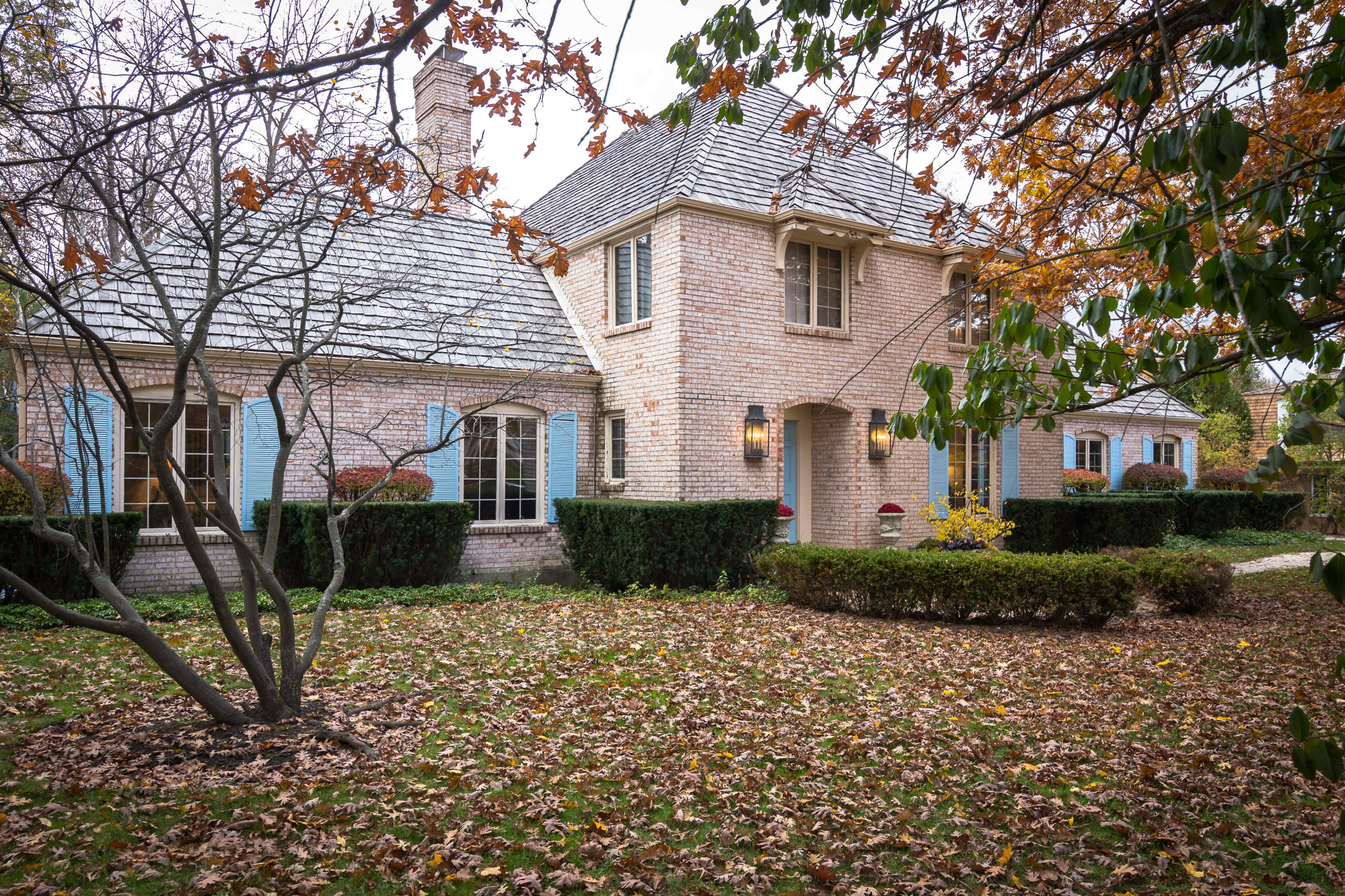 A brick house with blue shutters sits in a yard covered in autumn leaves, surrounded by neatly trimmed hedges and trees.