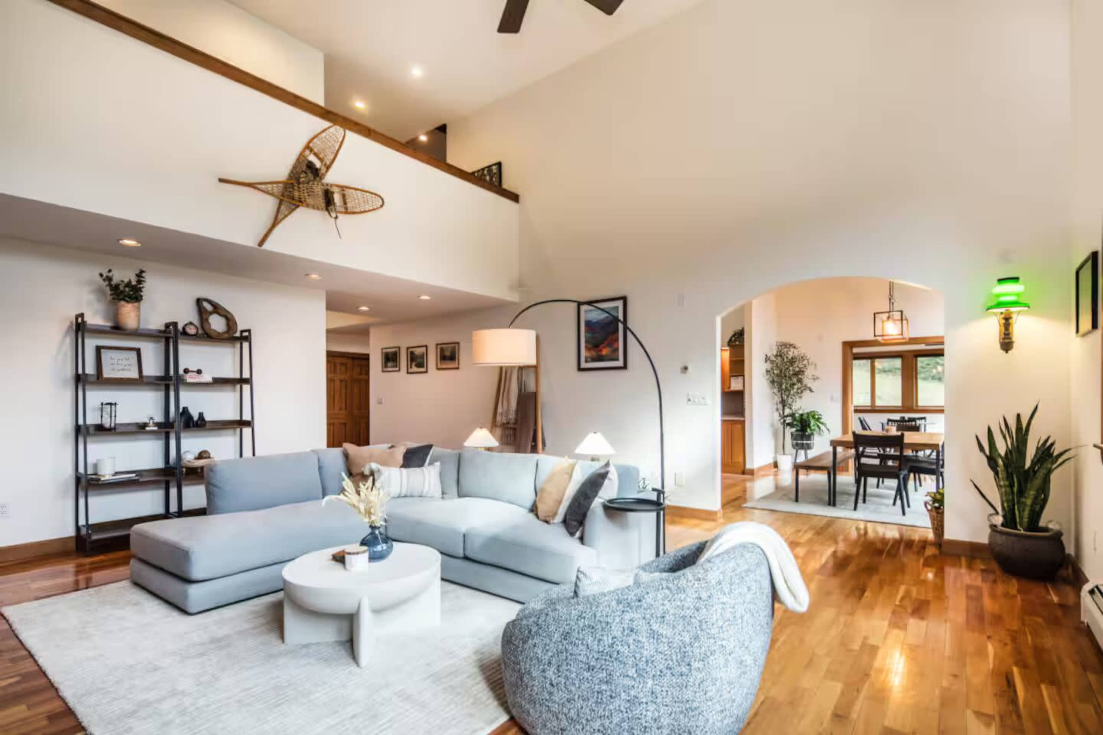 The image shows a spacious living area featuring a large sectional sofa, a round coffee table, and various decorative elements, with an open layout that connects to a dining area in the background.
