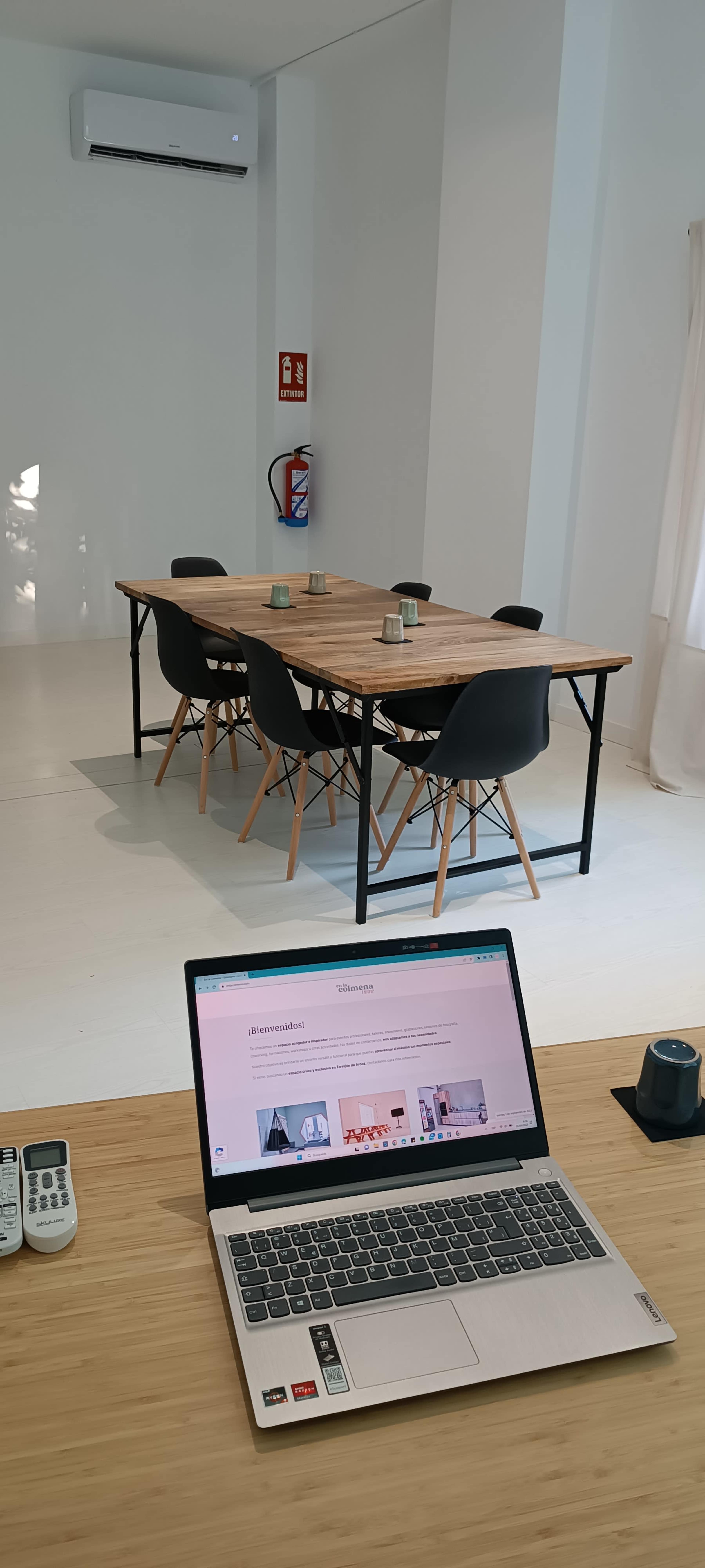 A wooden conference table with black chairs is positioned in a bright, minimalist room, with a laptop and various devices placed on a nearby wooden surface.