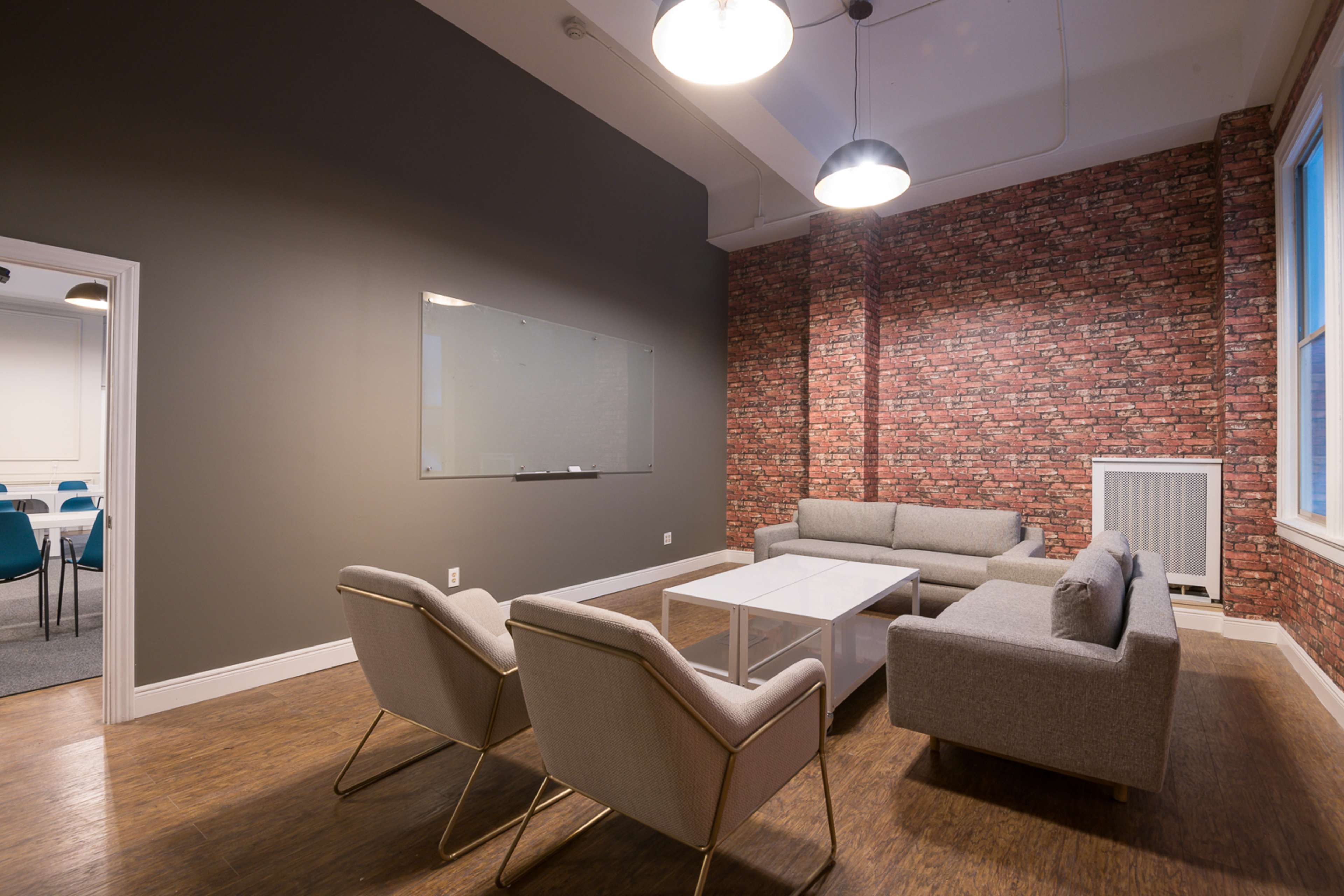 The image shows a modern lounge area with gray sofas and armchairs, a glass top coffee table, and a brick accent wall.