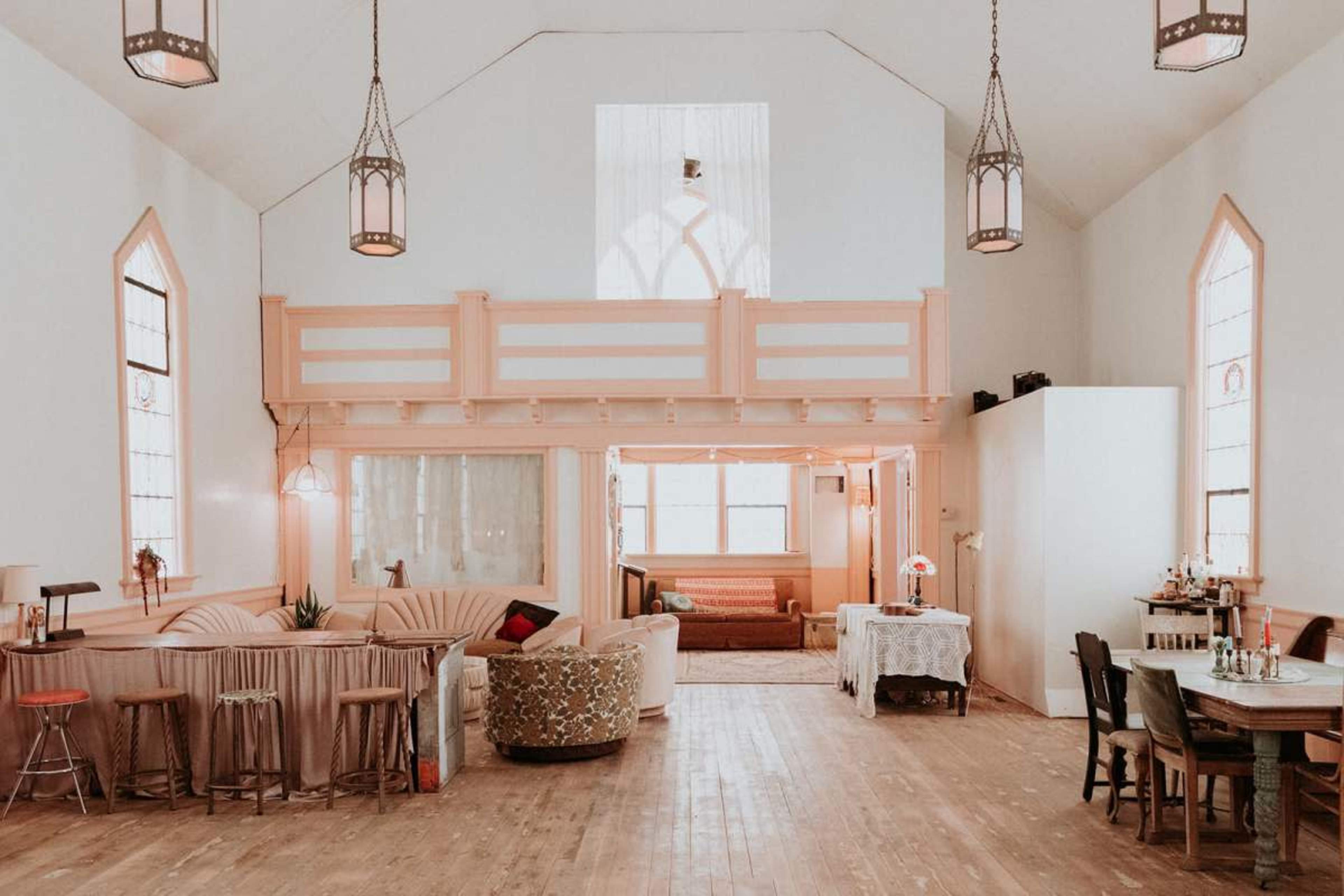 The image shows a spacious, well-lit interior of a room with high ceilings, featuring a mix of seating areas, wooden flooring, and various decorative elements.