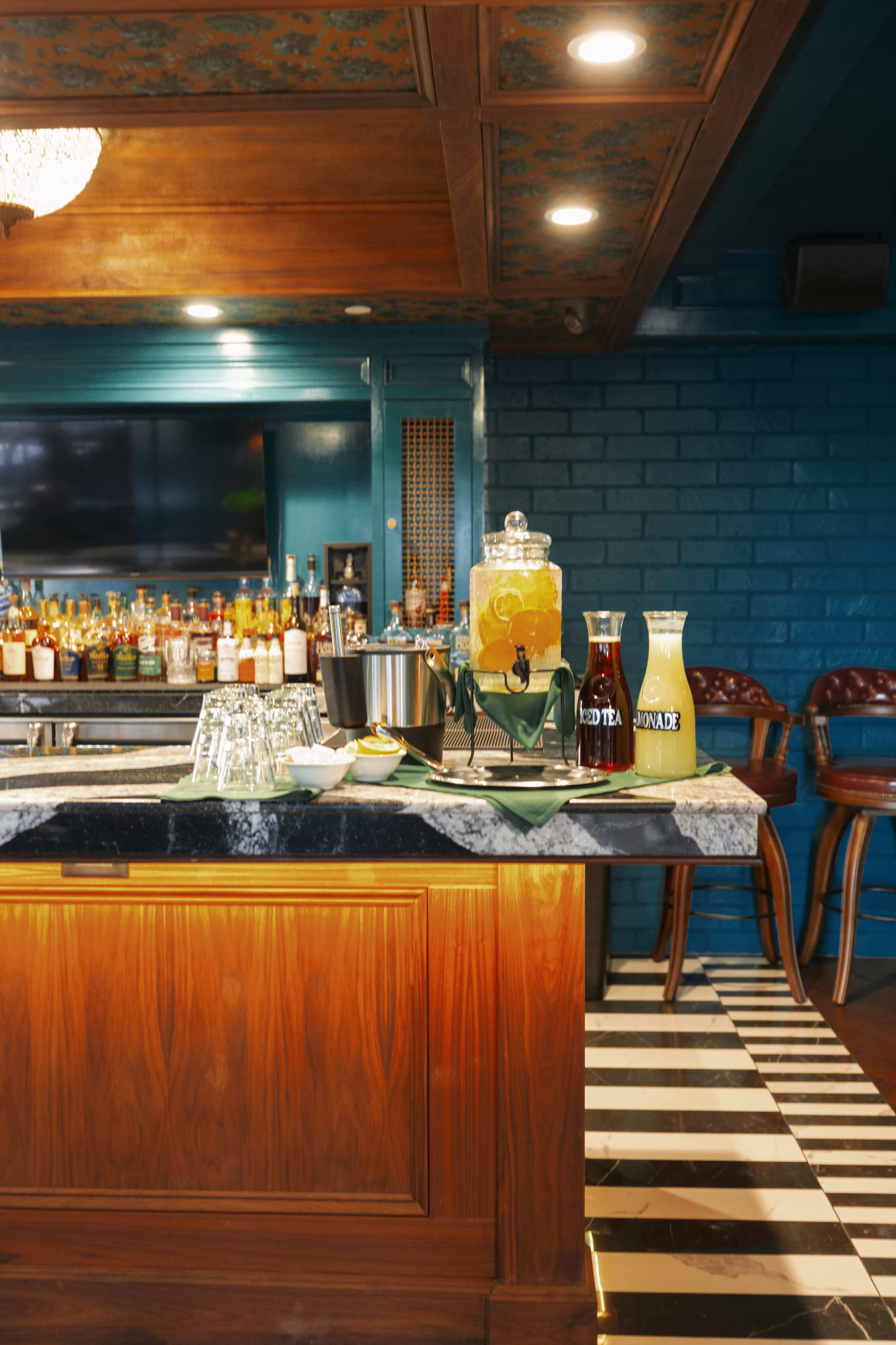 A polished bar area features a marble countertop, glass beverage dispensers, and a variety of liquor bottles displayed on shelves against a blue brick wall, with bar stools in the background.