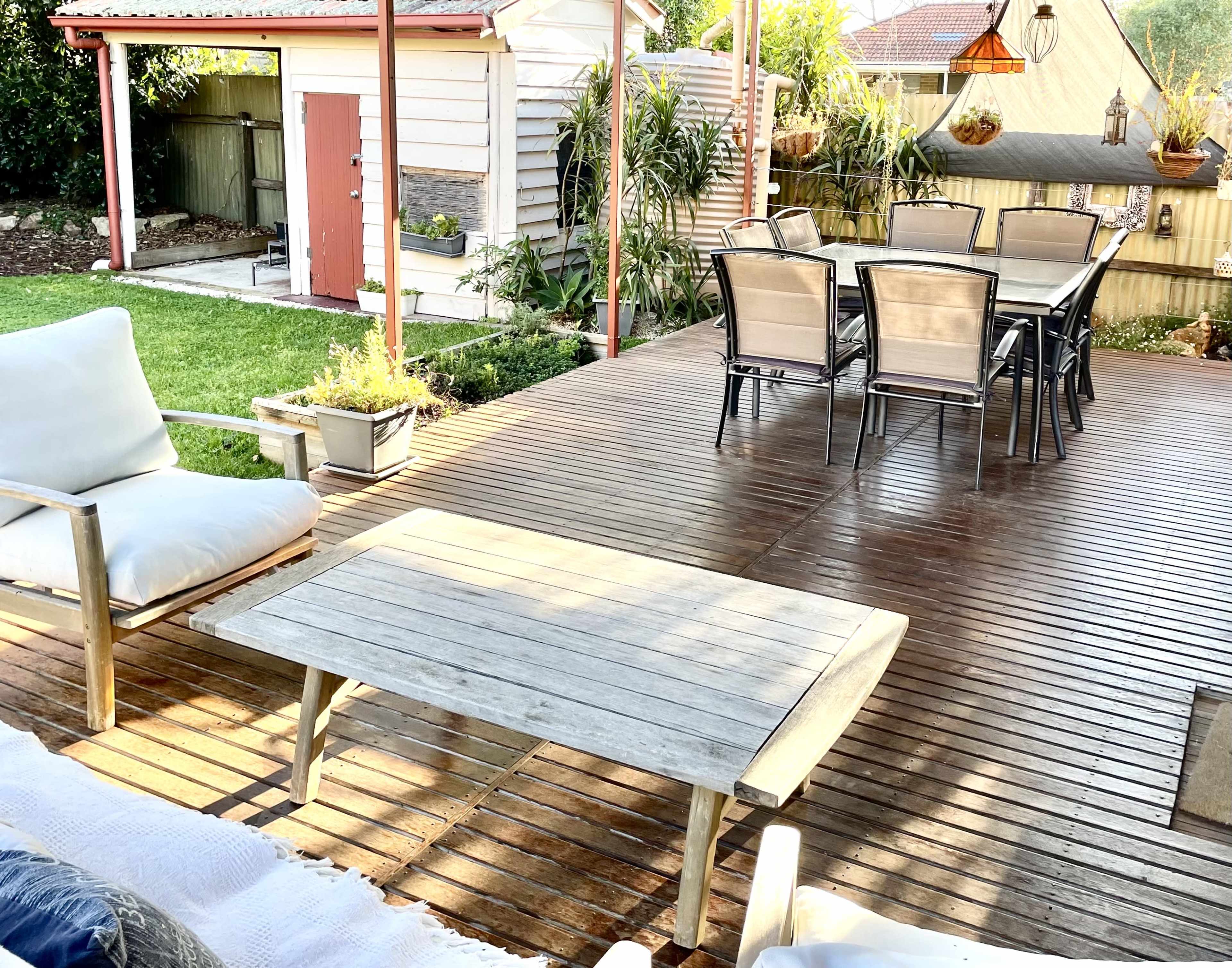 The image shows a wooden deck with a patio table and chairs, along with a lounge chair, surrounded by greenery and a shed in the background.