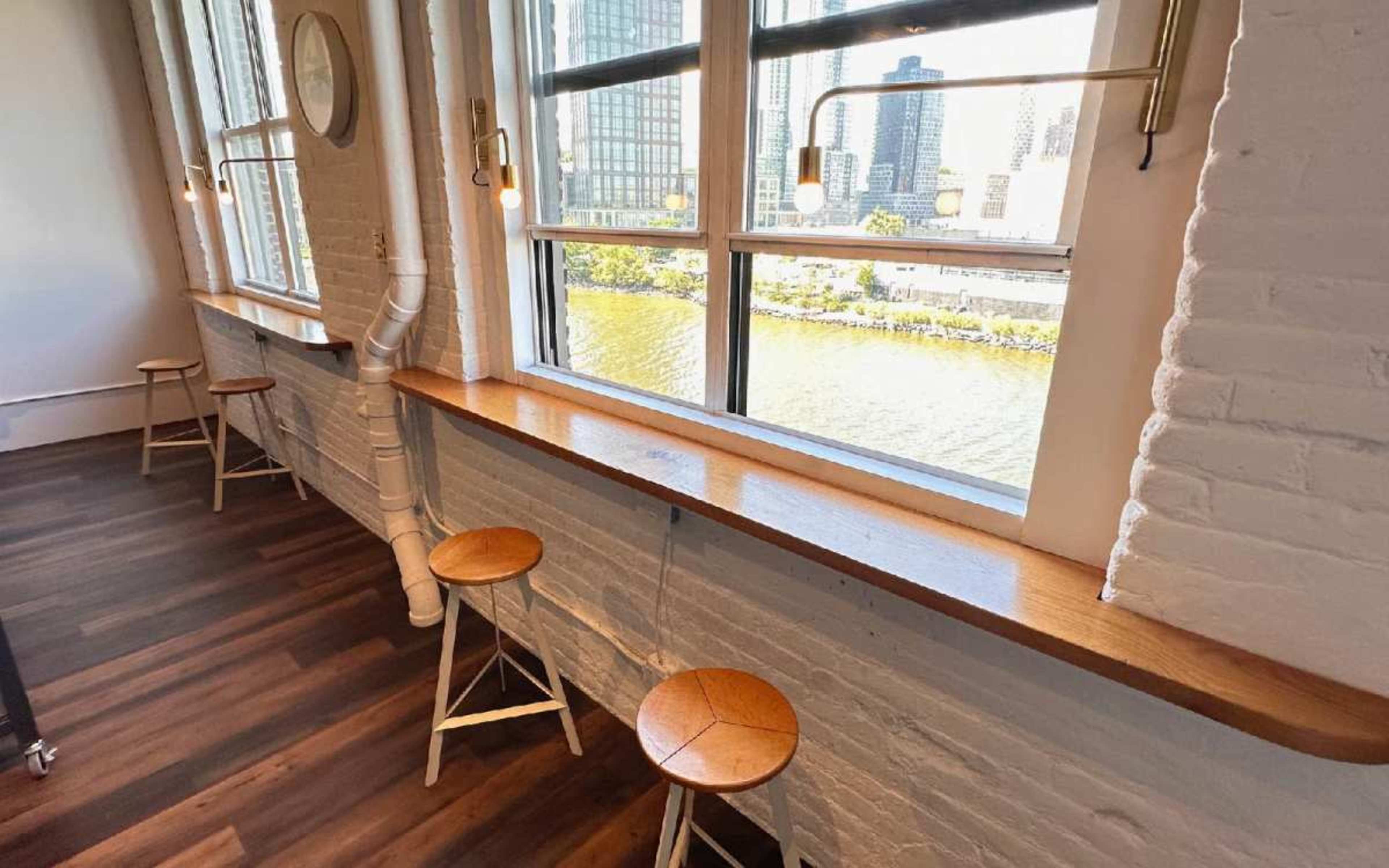 The image shows a light-filled room with large windows overlooking a river, featuring a wooden counter along the wall and three stools positioned beside it.