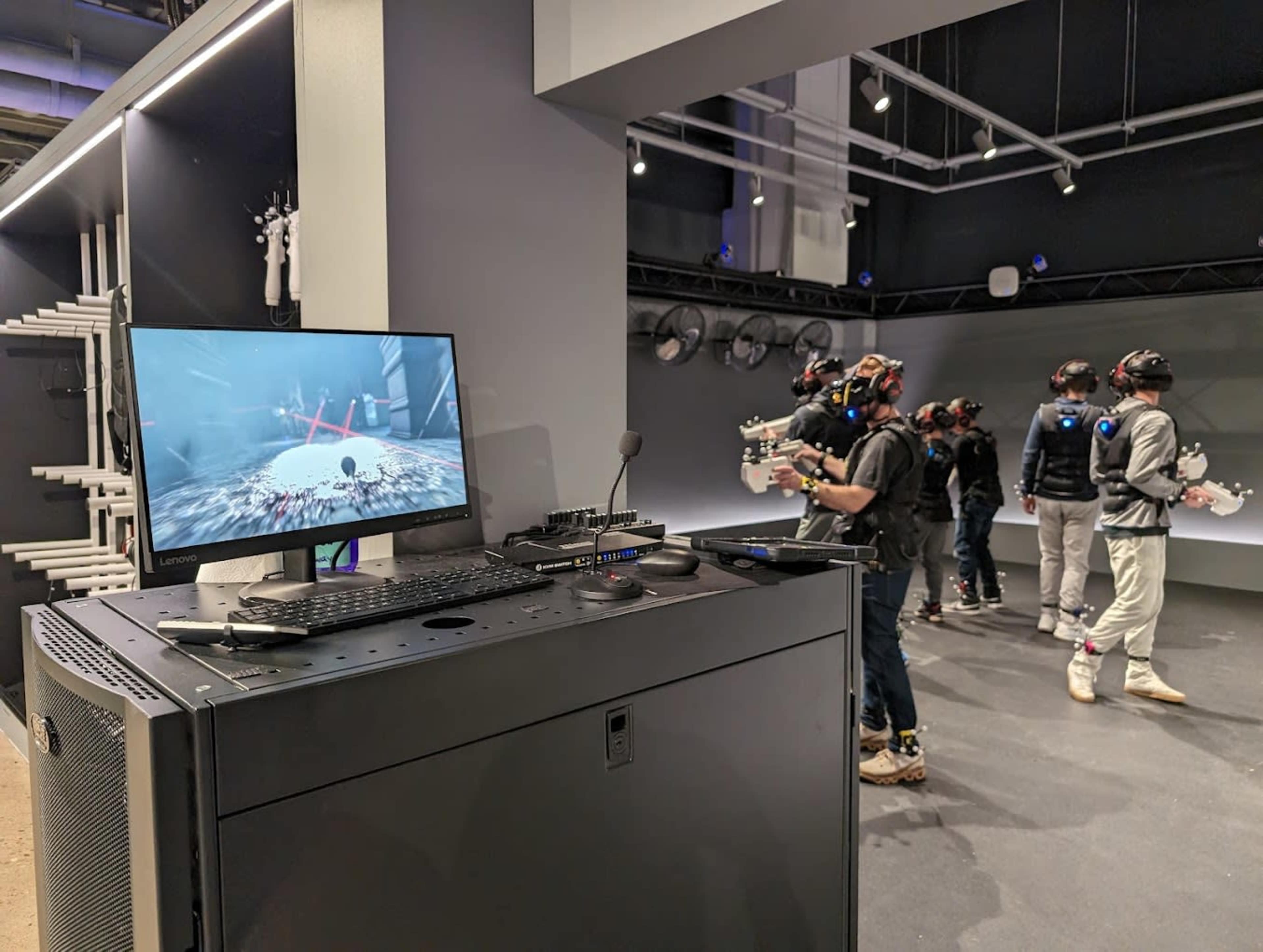 A group of individuals wearing virtual reality headsets and controllers stands in a spacious room equipped with screens and a central console displaying a virtual environment.