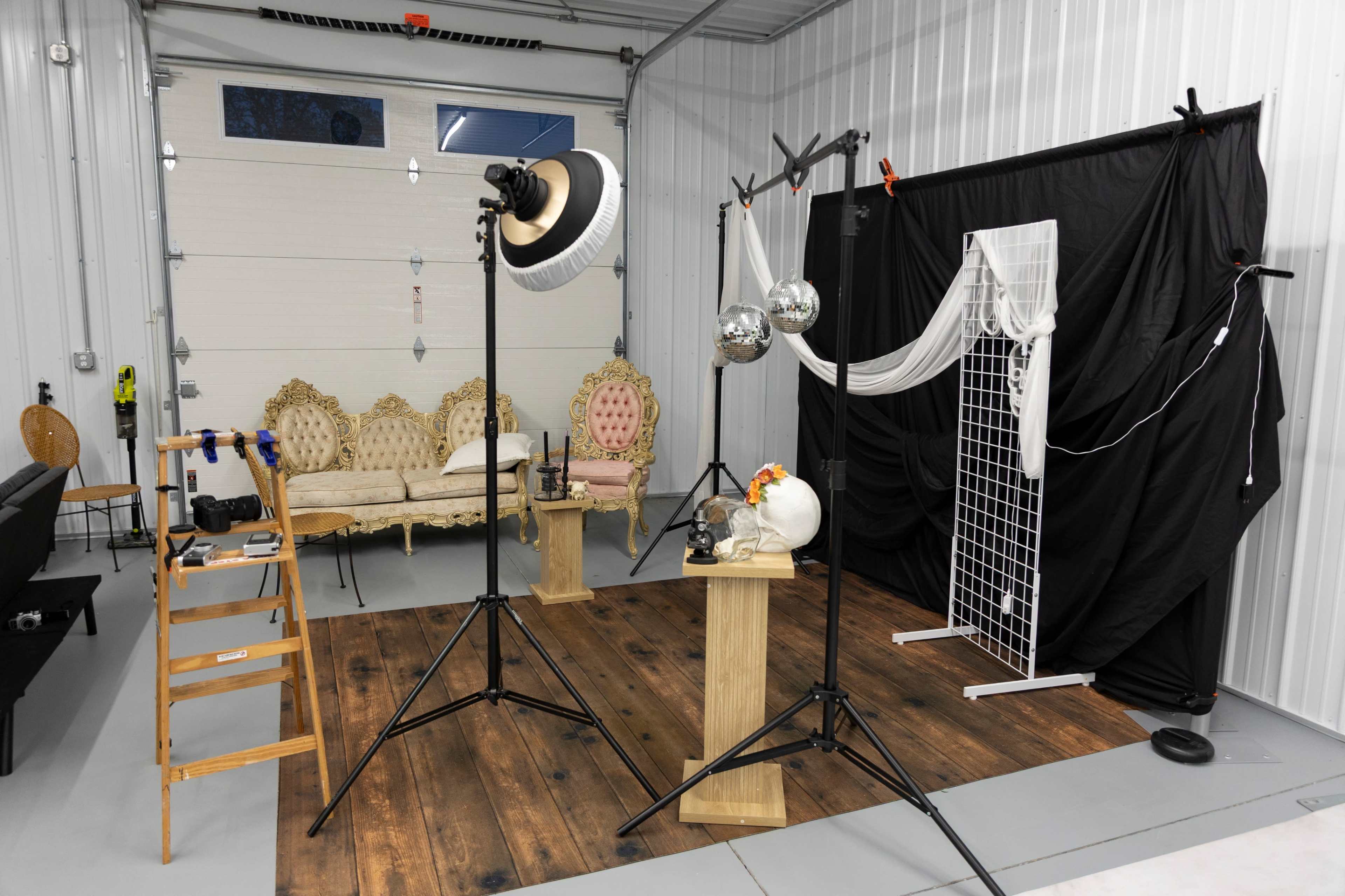 The image shows a photo studio setup with decorative furniture, lighting equipment, and a black backdrop draped with white fabric.