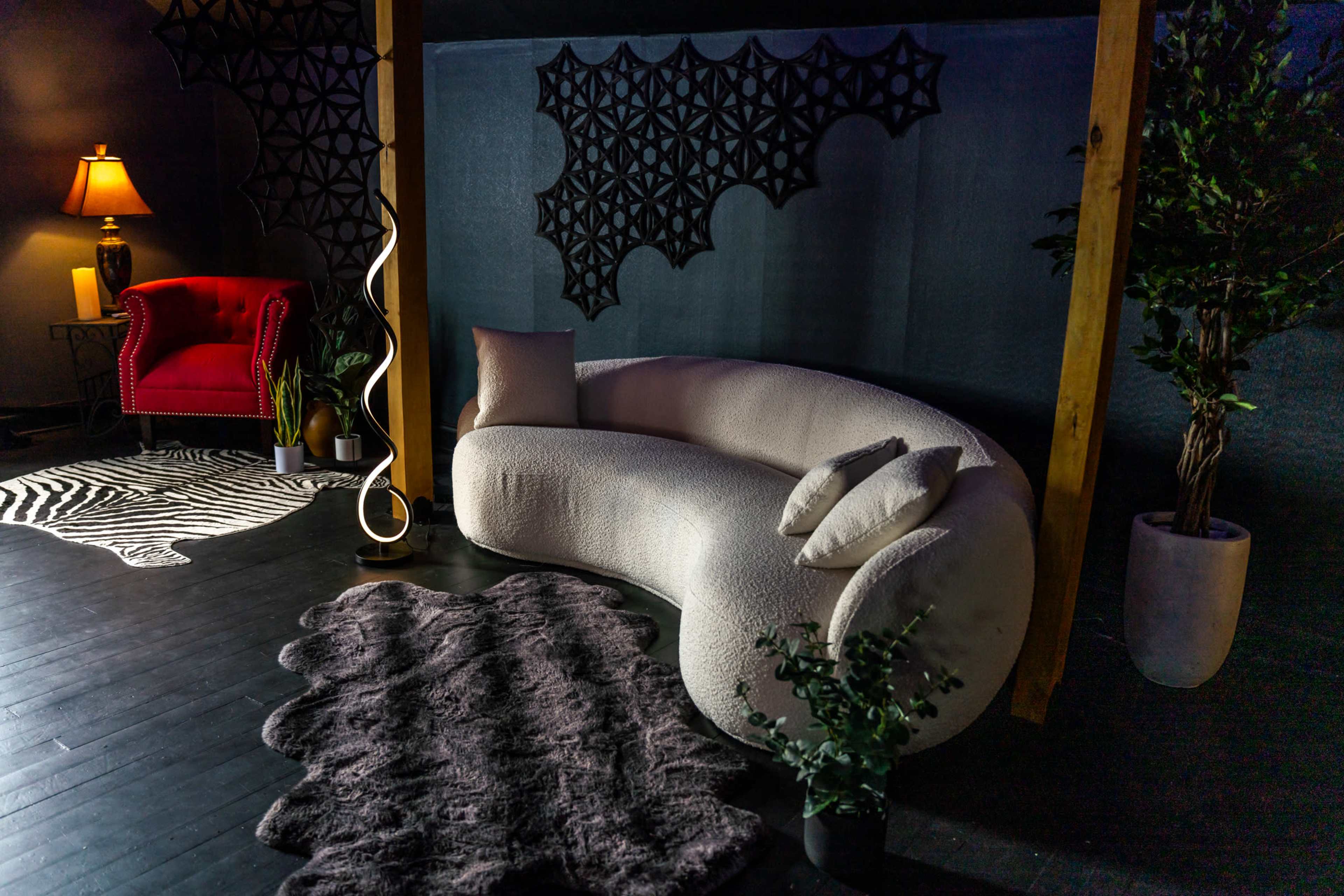 A modern living space featuring a curved white sofa, a red armchair, decorative plants, and various textured rugs against a dark backdrop.
