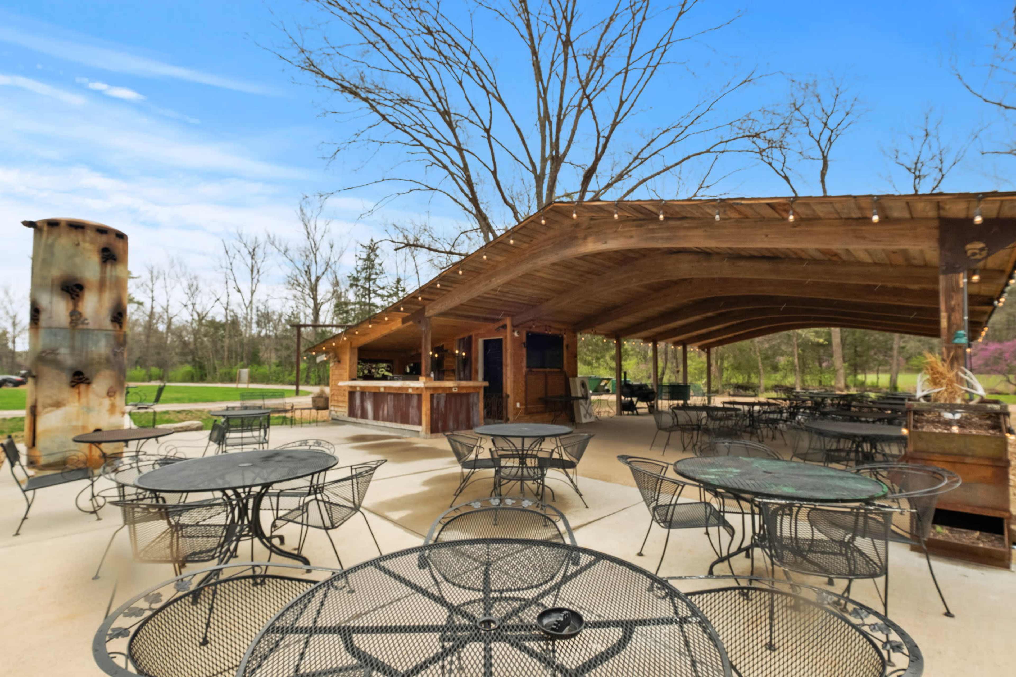 The image shows an outdoor dining area with metal tables and chairs under a large wooden canopy, surrounded by trees.