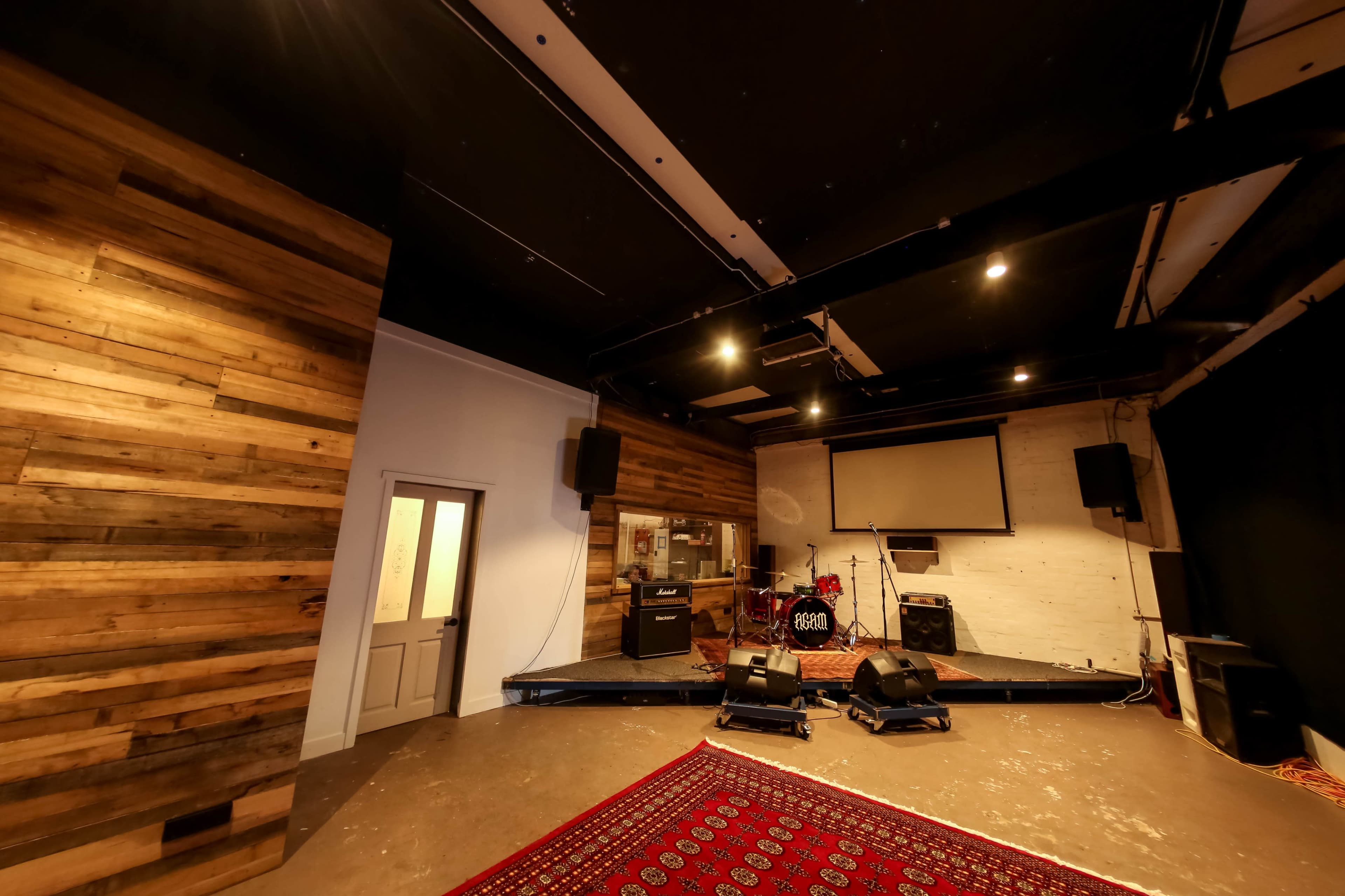 The image shows a music performance space with a wooden wall, a small stage, audio equipment, and a red patterned rug on the floor.