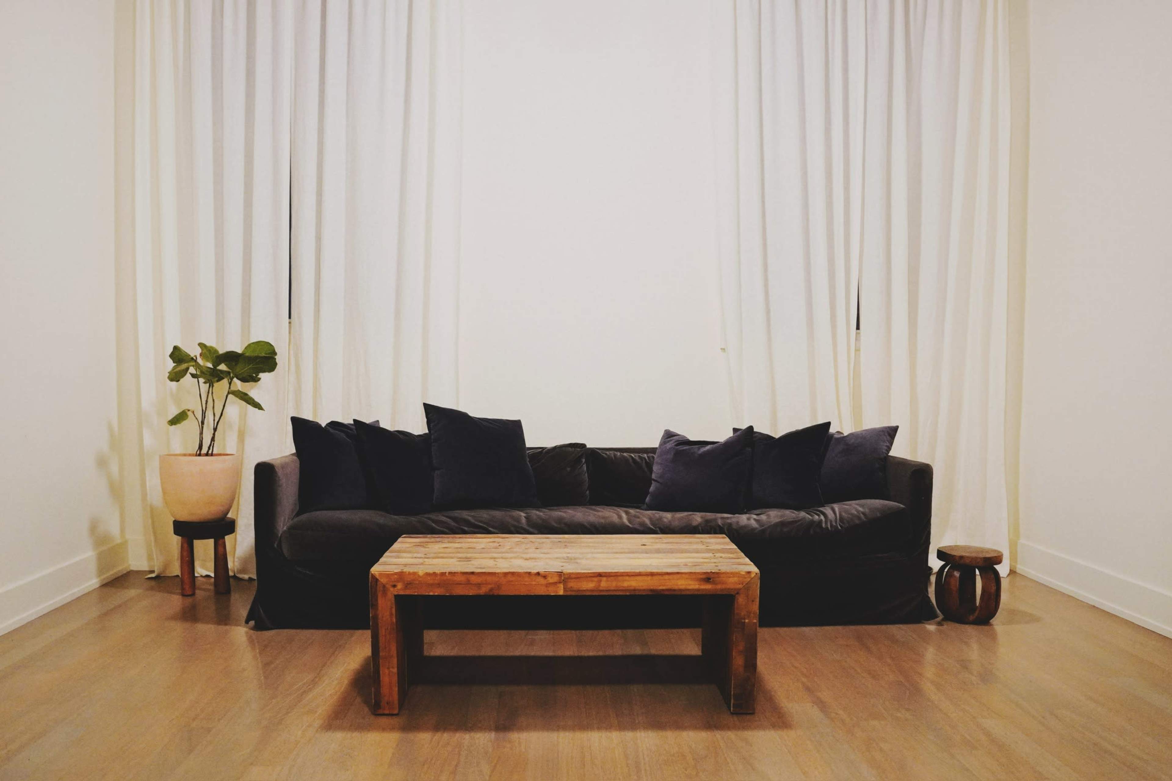 A dark couch with several cushions sits against a wall beneath large curtains, accompanied by a wooden coffee table and a potted plant.