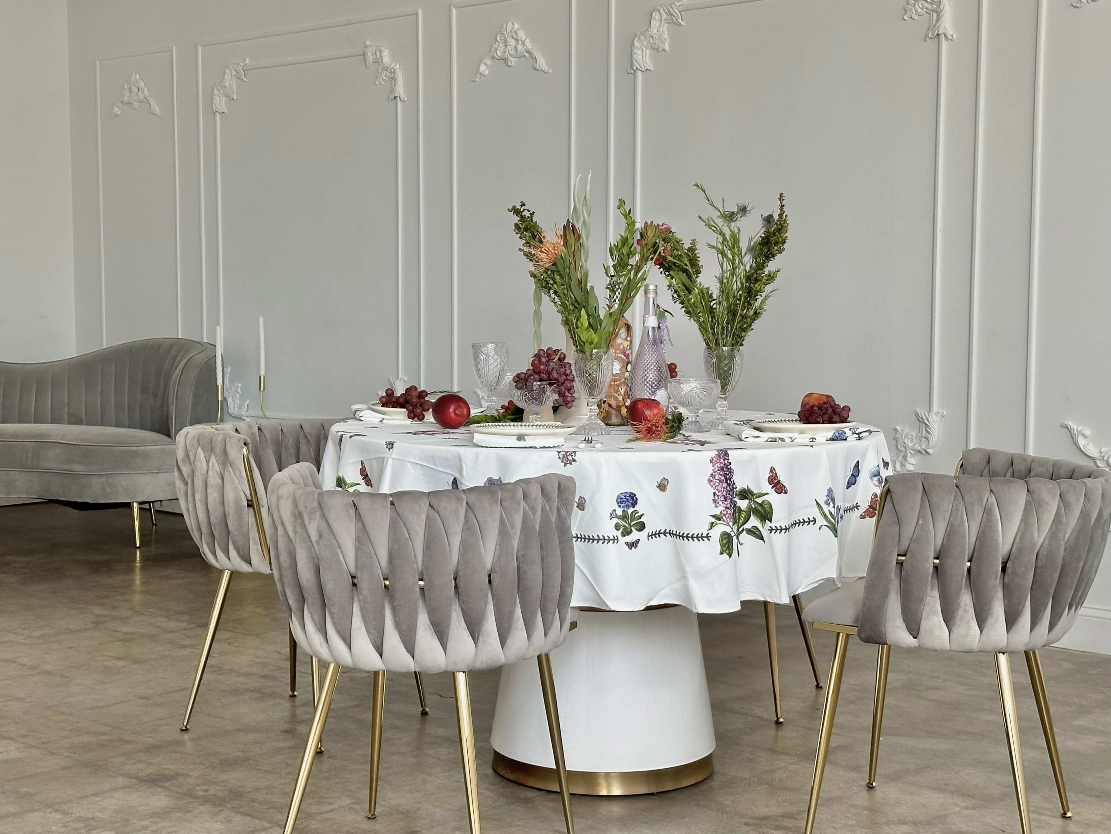 A round dining table with a floral tablecloth is set with various foods and decorative plants in a stylish room featuring a minimalist design.