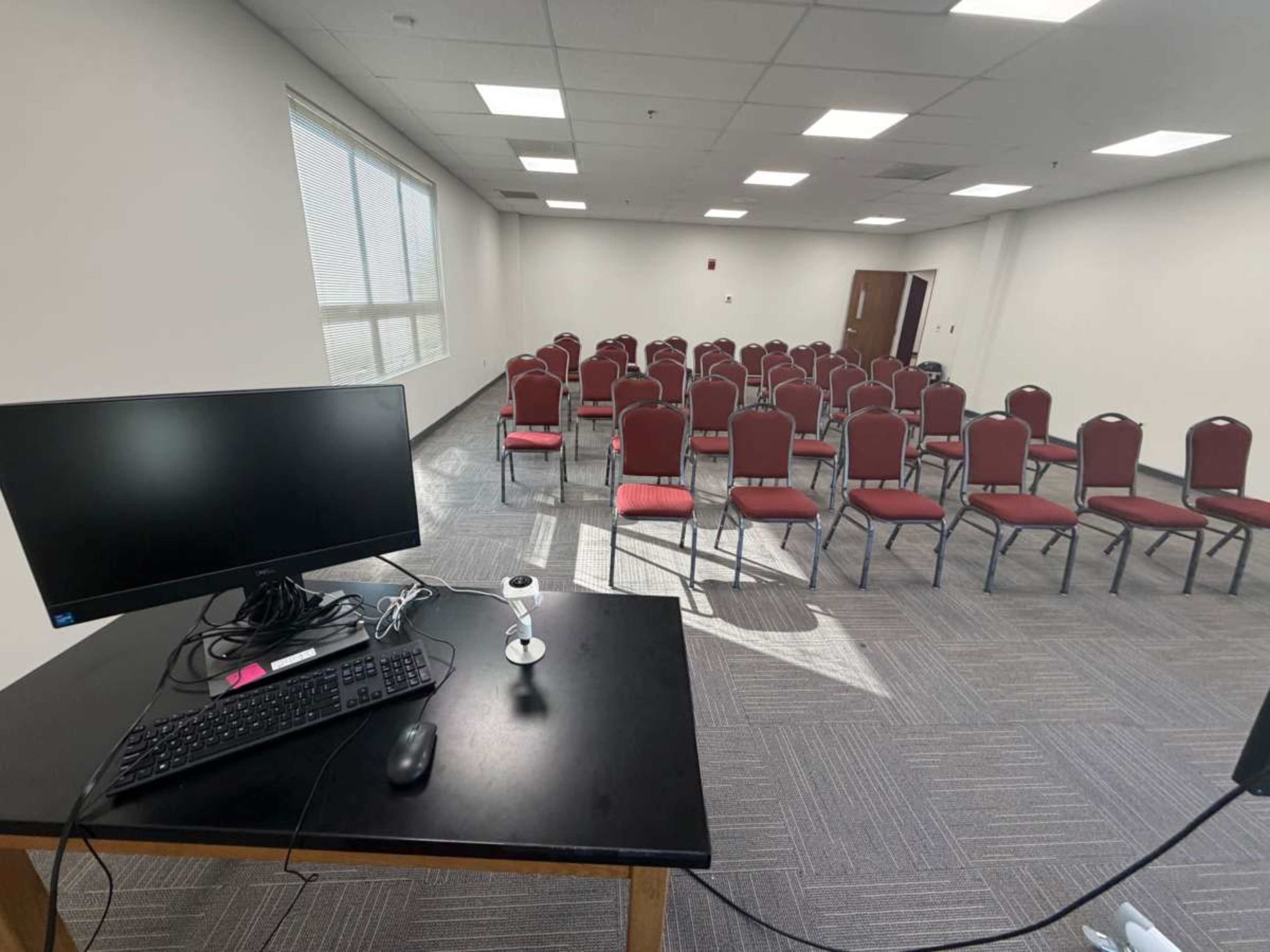 The image shows a room set up with rows of red chairs facing a table containing a computer and camera.