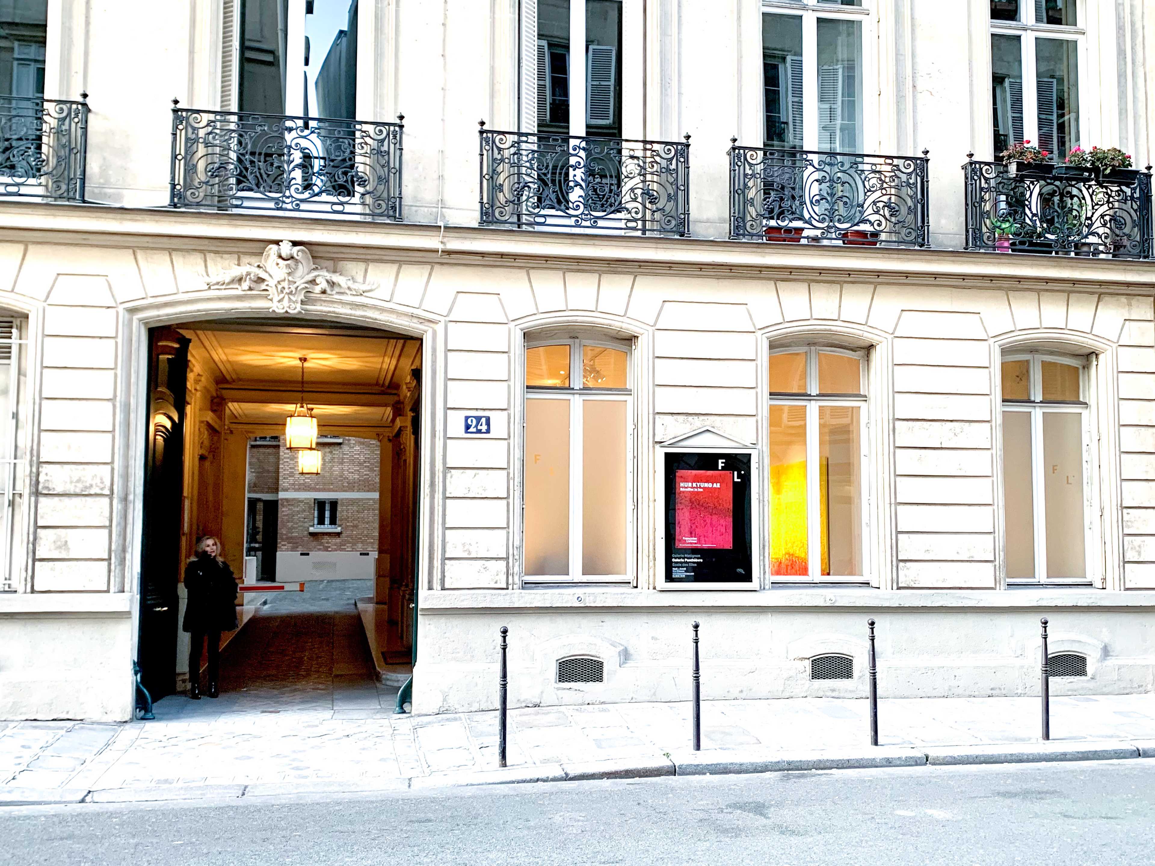 A person stands outside a building with tall windows and intricate ironwork, located on a quiet street.