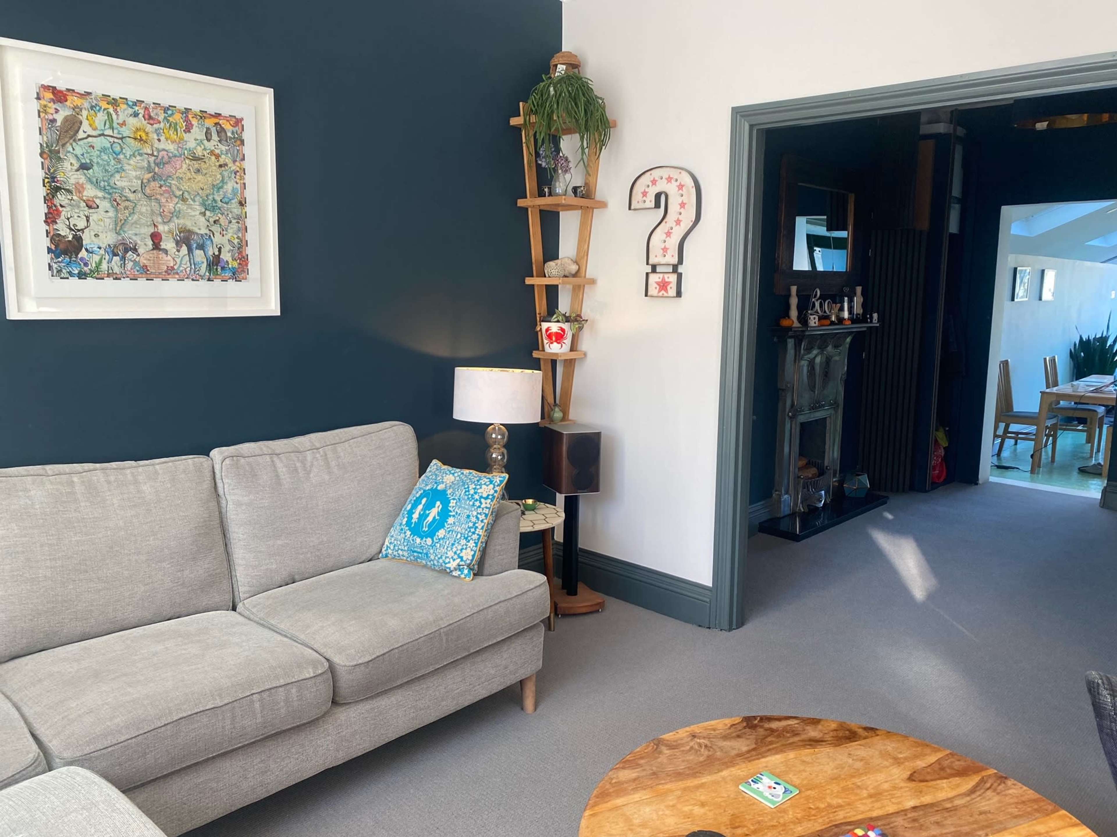 The image shows a living room with a gray sofa, a decorative wall hanging, a wooden ladder shelf with plants, and an adjacent doorway leading to a dining area.