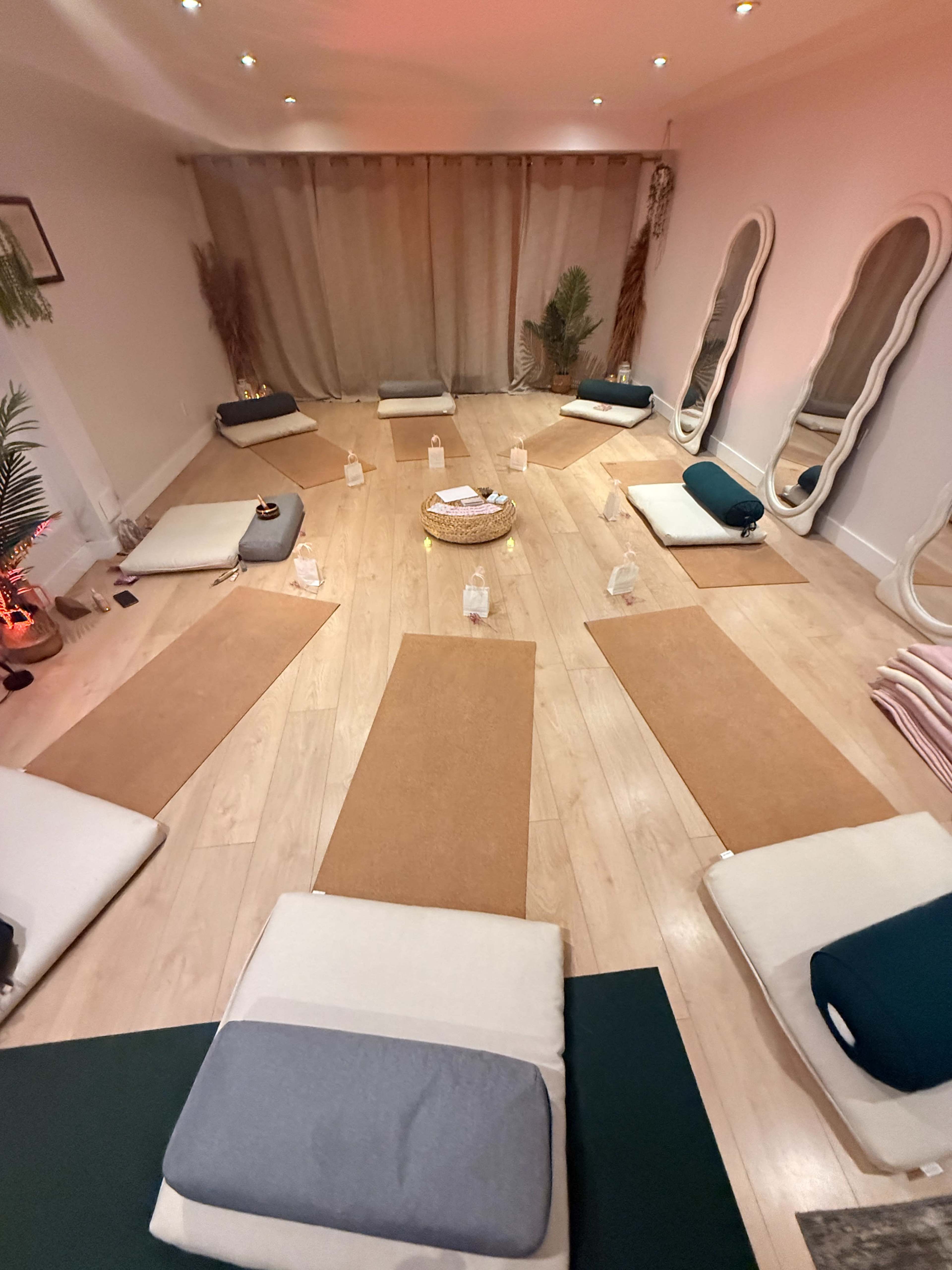 A serene wellness studio is set up with mats, cushions, and water bottles arranged in a circular layout, surrounded by soft lighting and curtains.