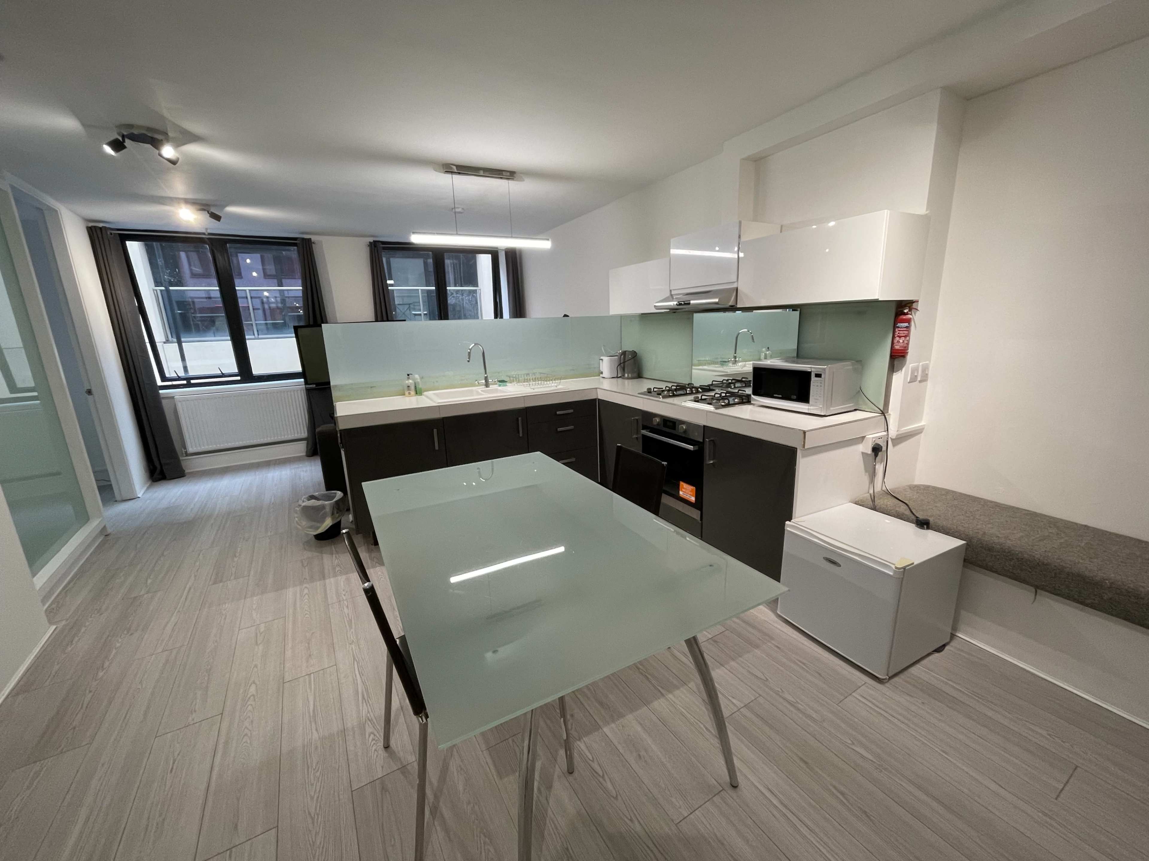 The image shows a modern kitchen with a glass dining table, a gas stove, a microwave, and large windows allowing natural light.