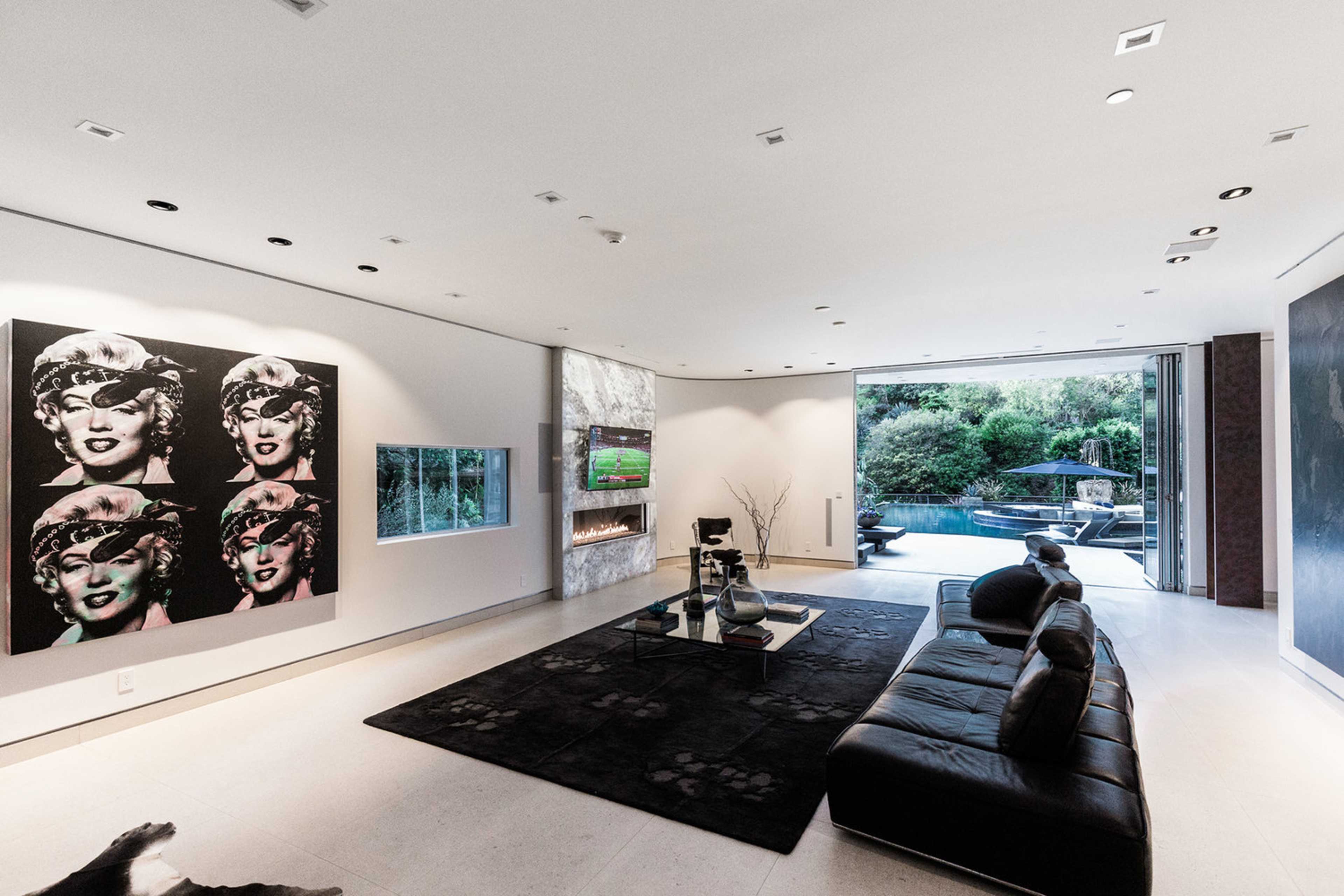 A modern living room features a large black sofa, a striking Marilyn Monroe art piece on the wall, and an expansive view of the pool area through open sliding glass doors.