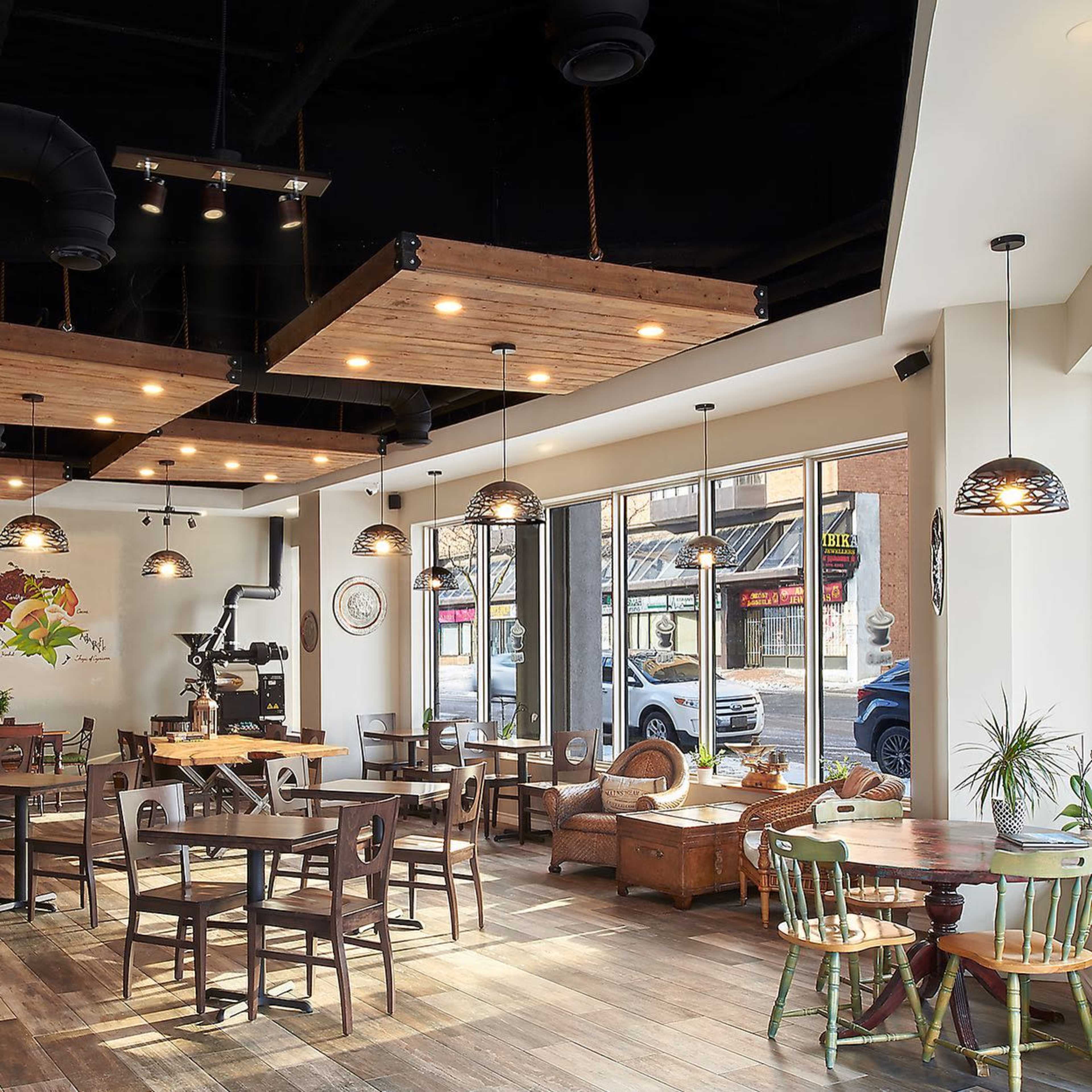 A modern restaurant with wooden decor, tables, and chairs, illuminated by pendant lights, and large windows overlooking a street.