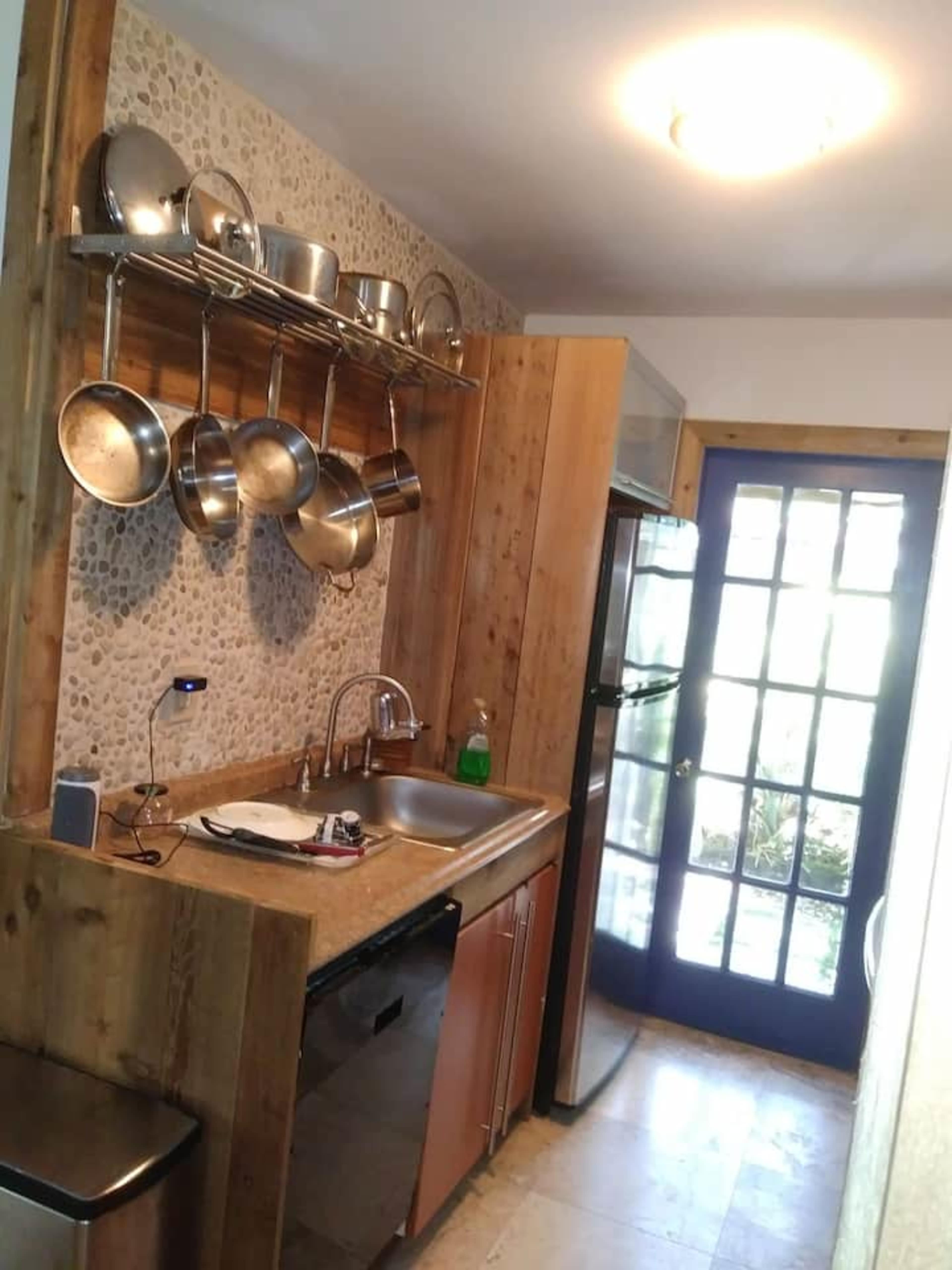 The image shows a small kitchen area with a stainless steel sink, hanging pots, and a doorway leading to an outdoor space.