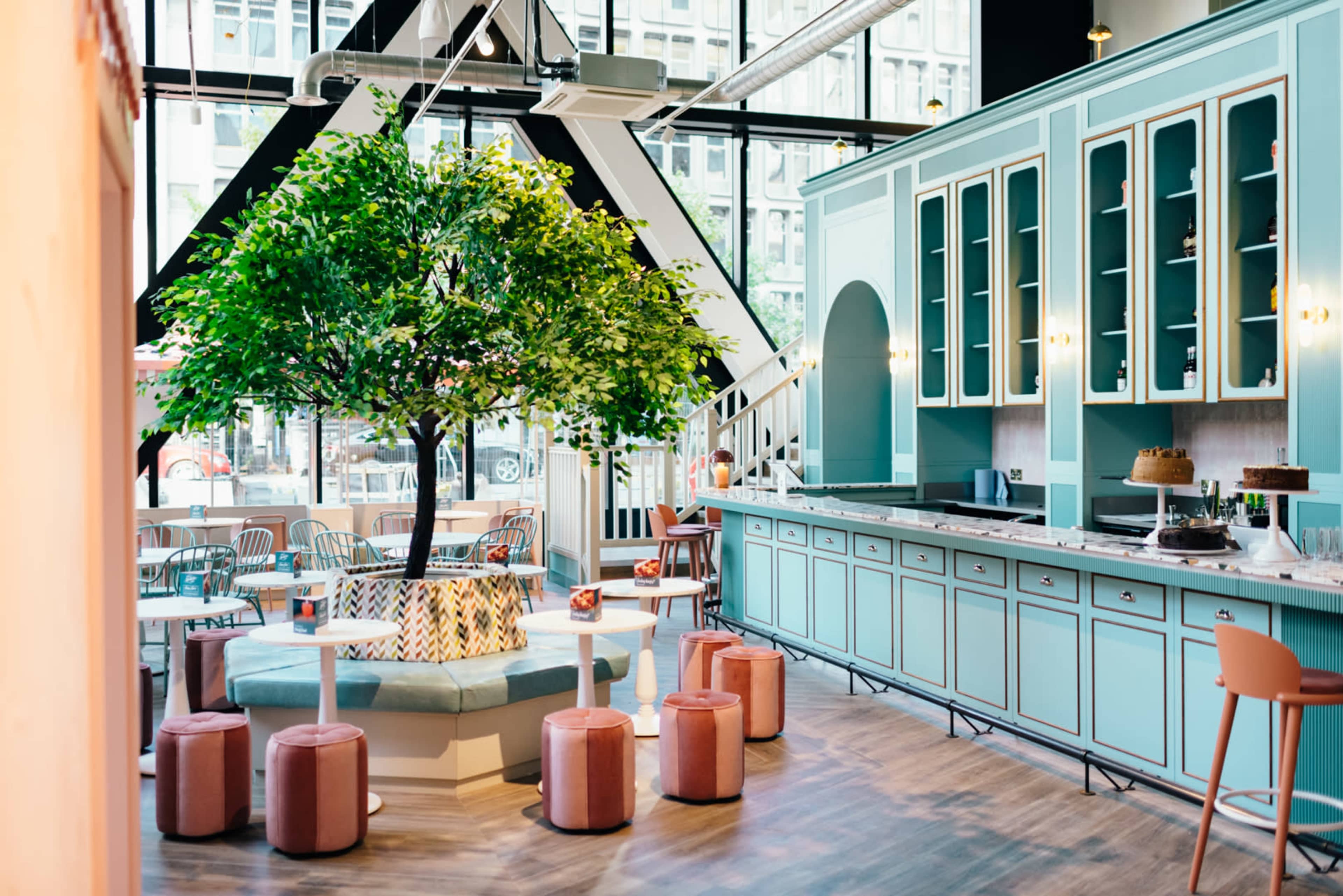 A modern café features a large artificial tree in the center, surrounded by pastel-colored seating, with a light blue kitchen area in the background.