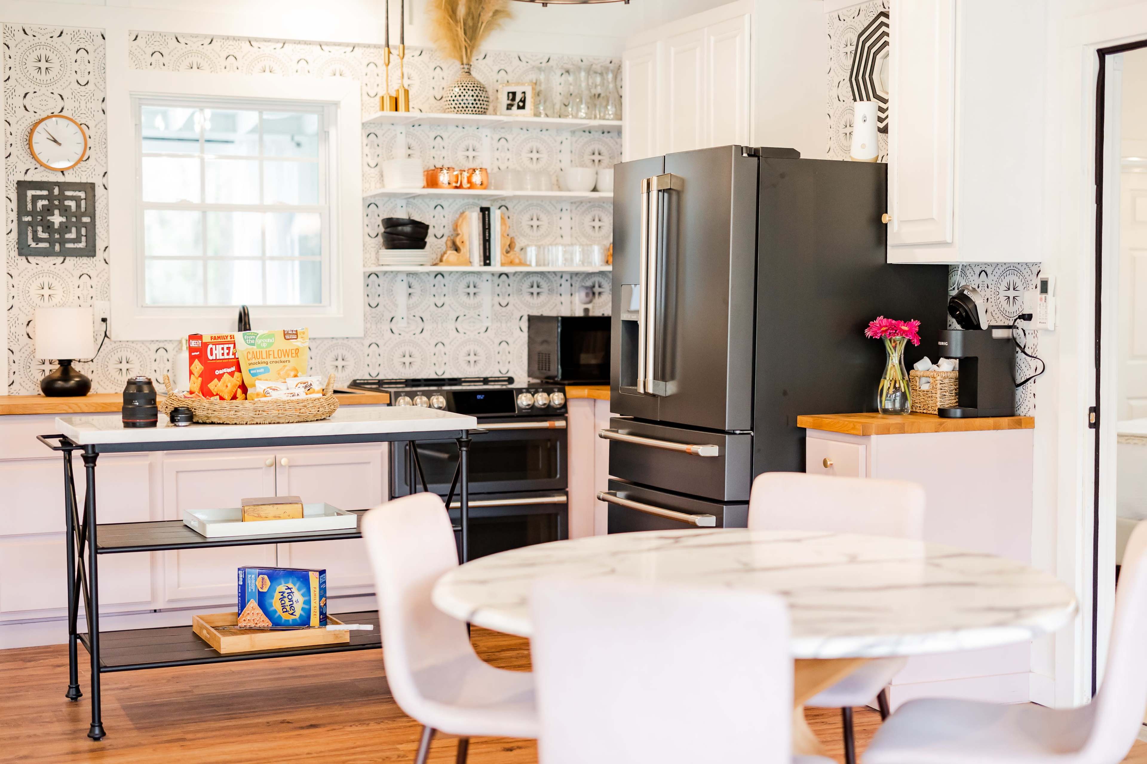 The kitchen features a black refrigerator, white cabinetry, and a round marble dining table, with decorative elements on the walls and a vase of flowers on the counter.