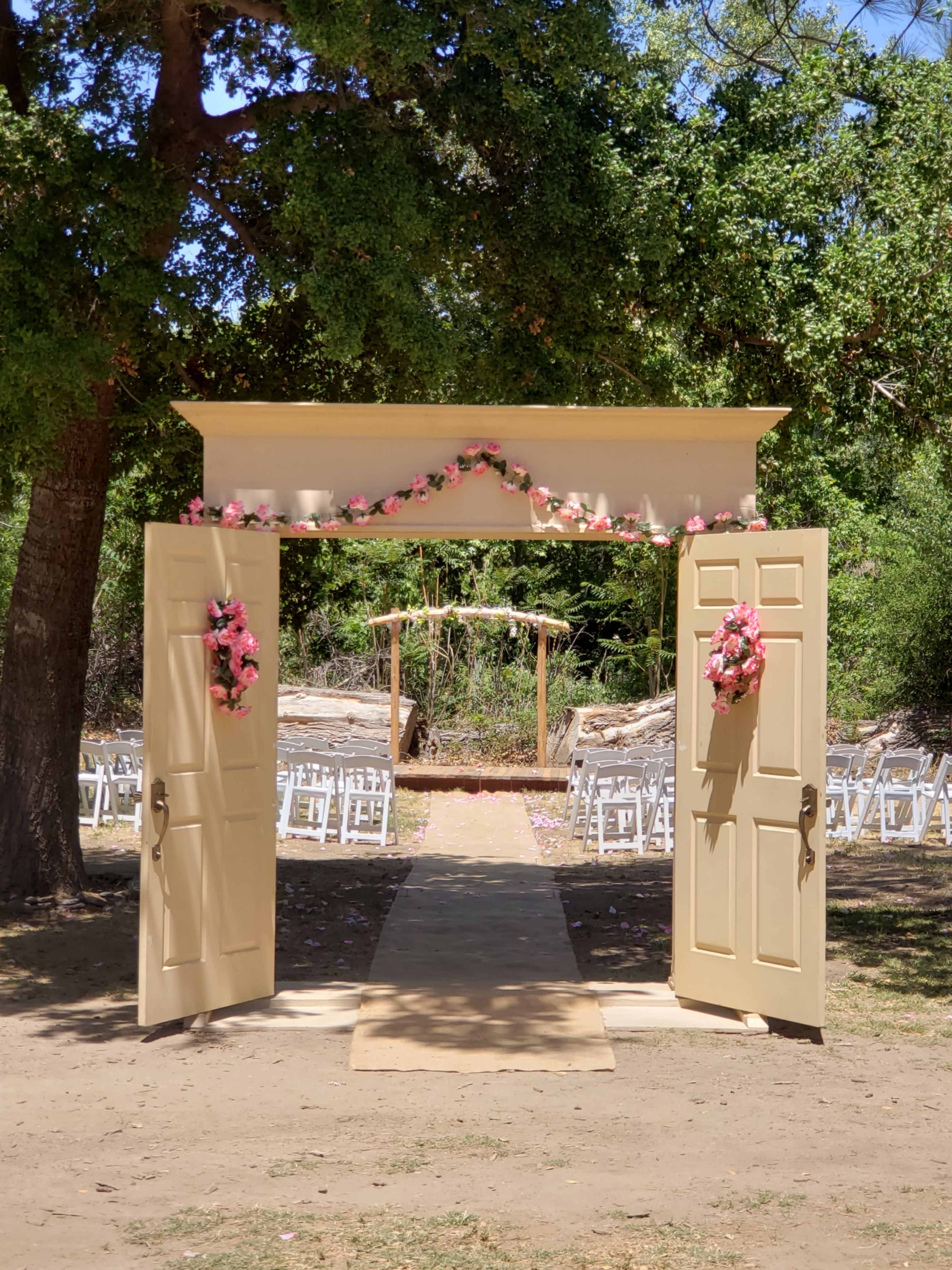 An open double door adorned with pink flowers leads to an outdoor wedding ceremony space featuring chairs and a decorative arch.