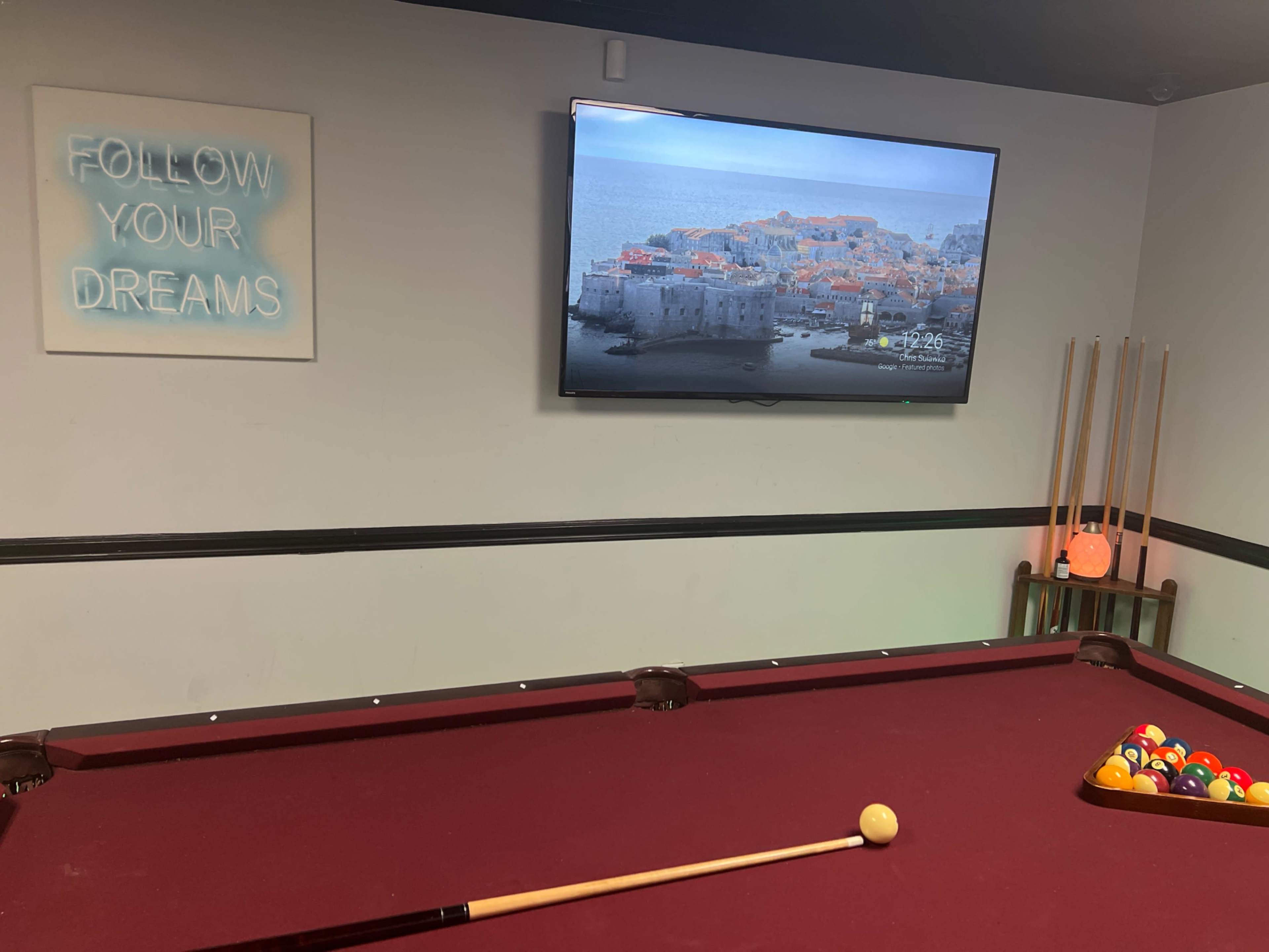 The image shows a game room with a red pool table, a television displaying a coastal scene, and a wall art piece that says "FOLLOW YOUR DREAMS."
