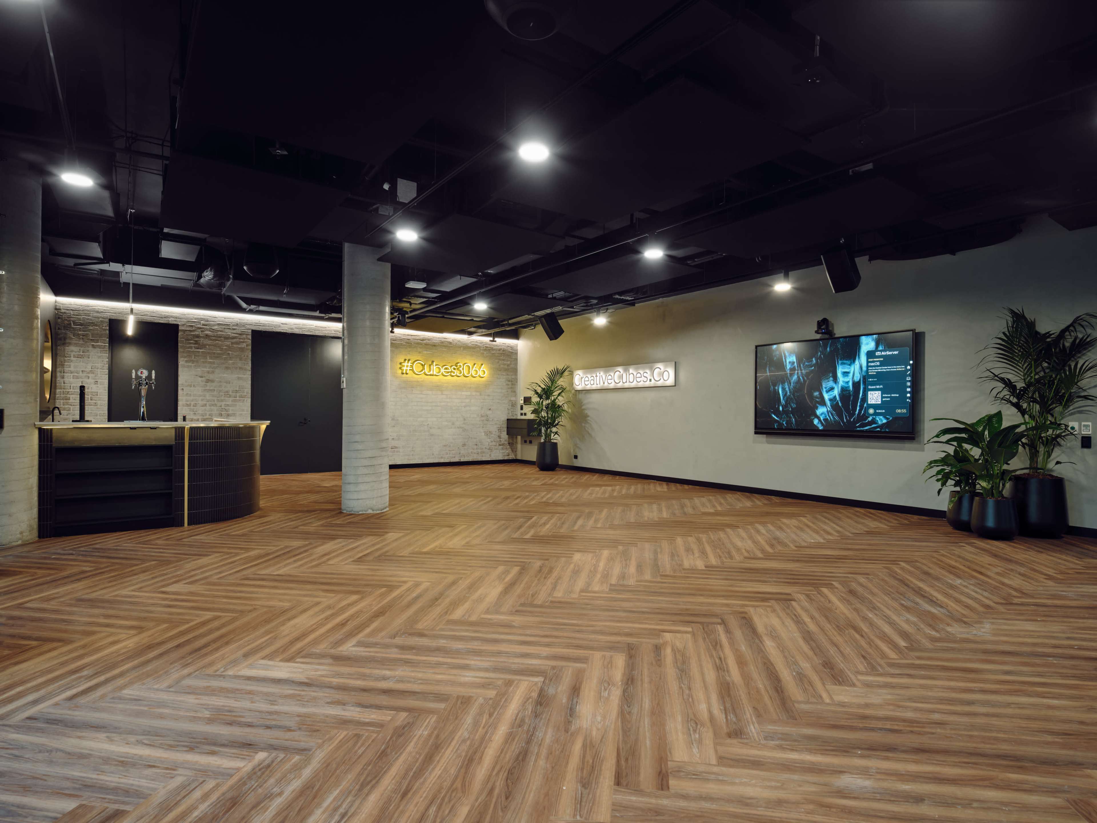 The image shows a modern, spacious lobby area with wooden flooring, a reception desk, and a large screen displaying digital content on the wall.