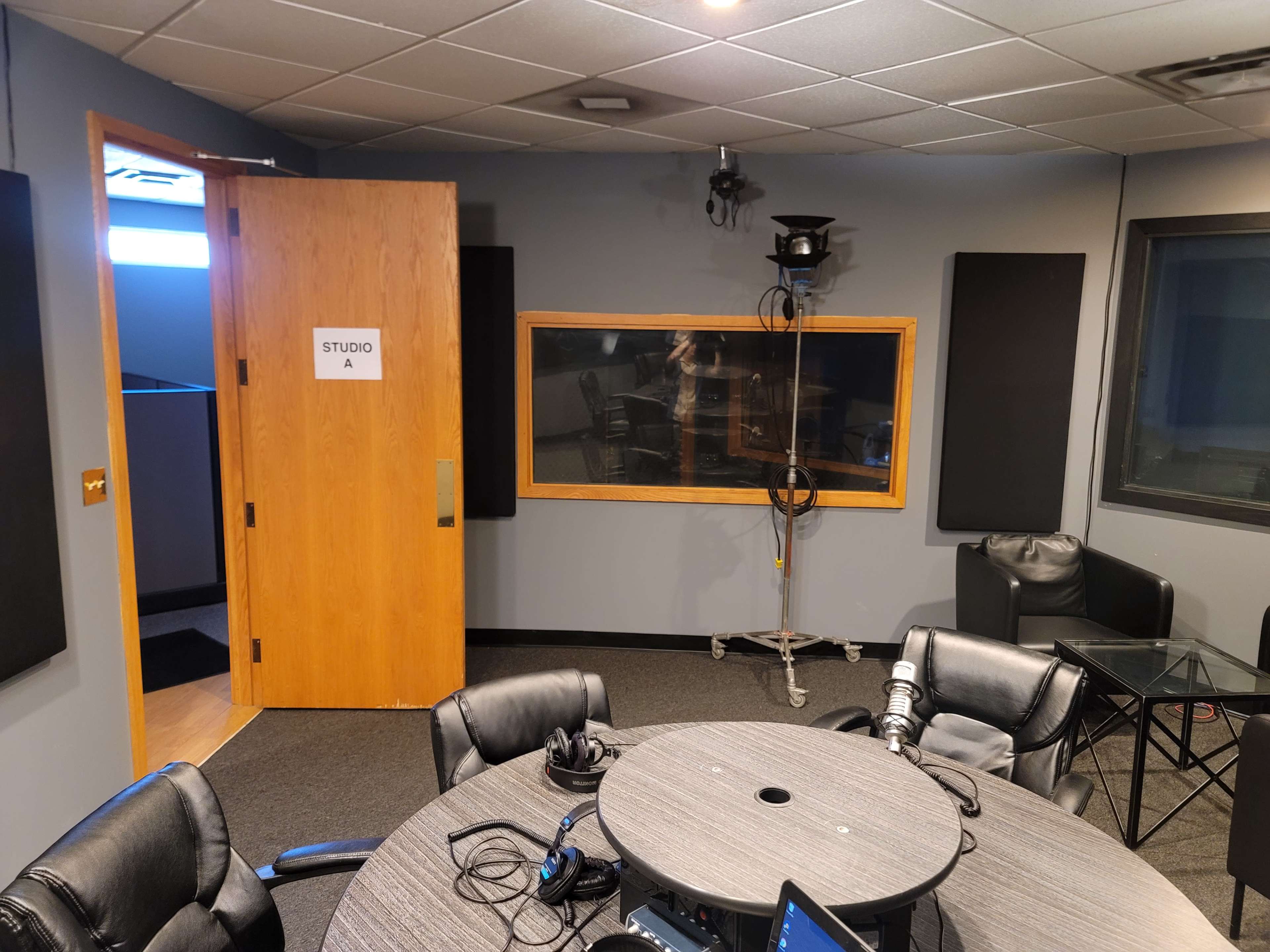 A recording studio with a wooden door labeled "STUDIO A," a circular table surrounded by black chairs, and soundproofing panels on the walls.