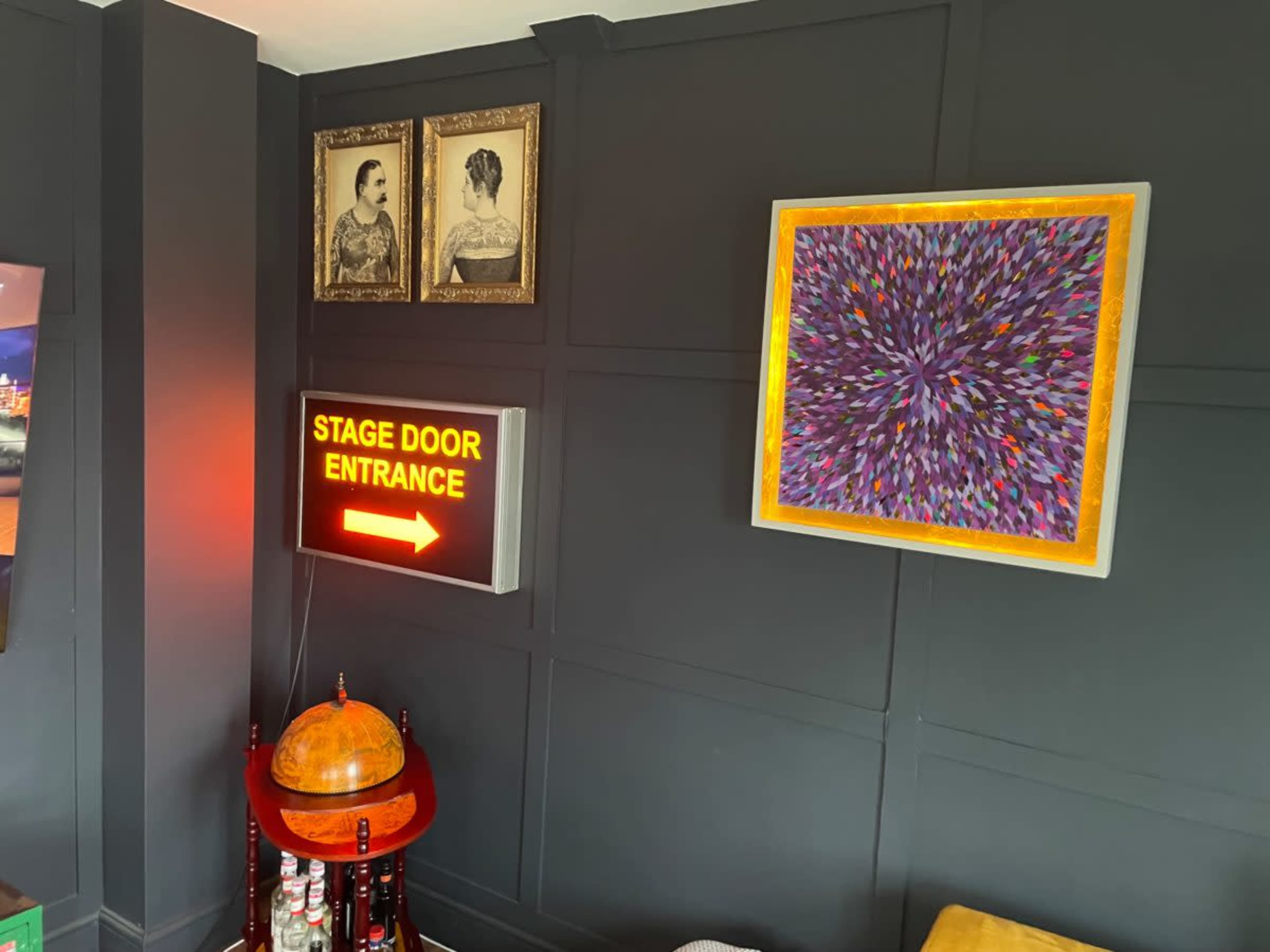 The image shows a dark room with a lighted sign that says "STAGE DOOR ENTRANCE" pointing to the right, surrounded by framed artworks and a globe on a stand.