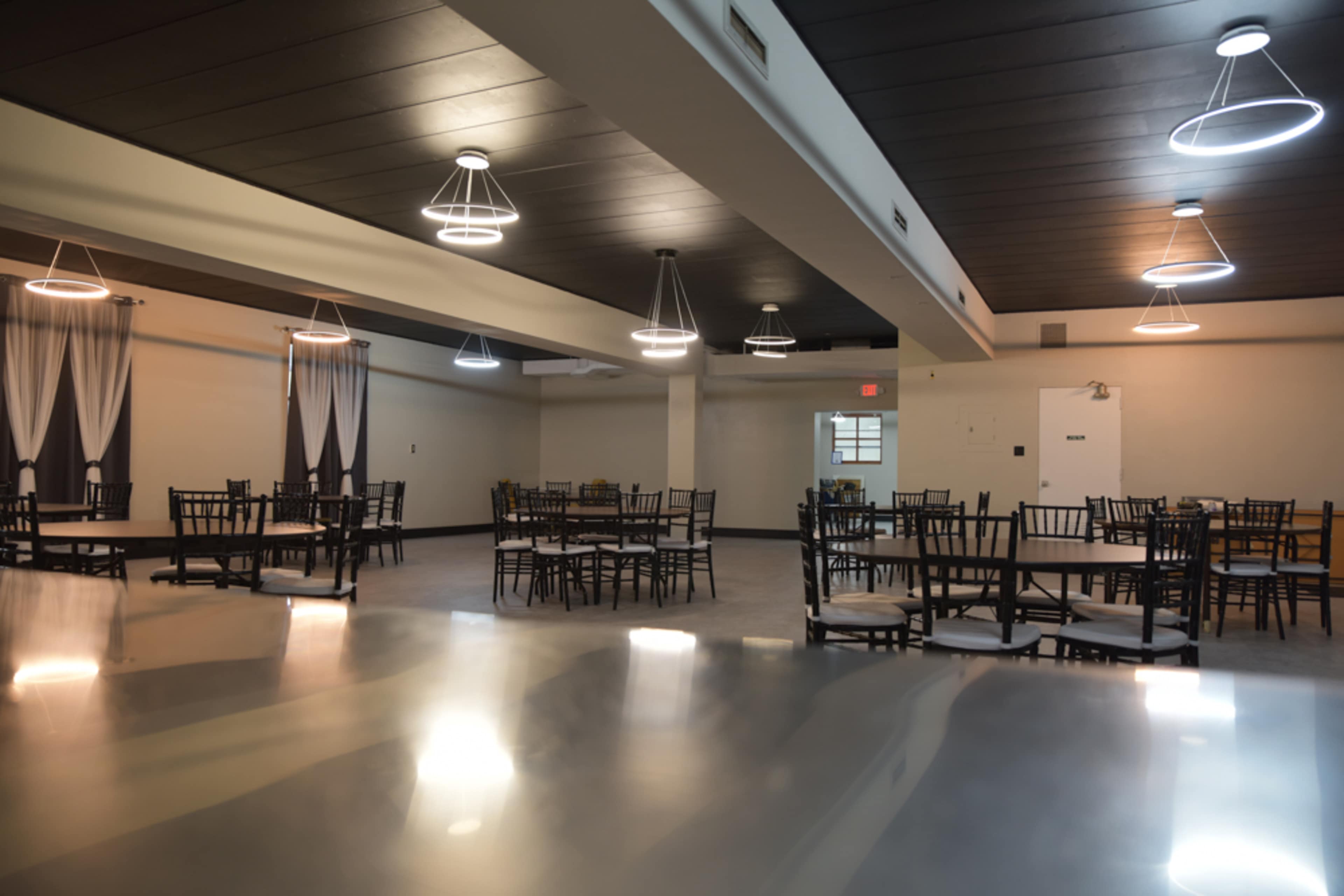 An empty banquet hall with several round tables and chairs arranged, illuminated by modern pendant lighting.