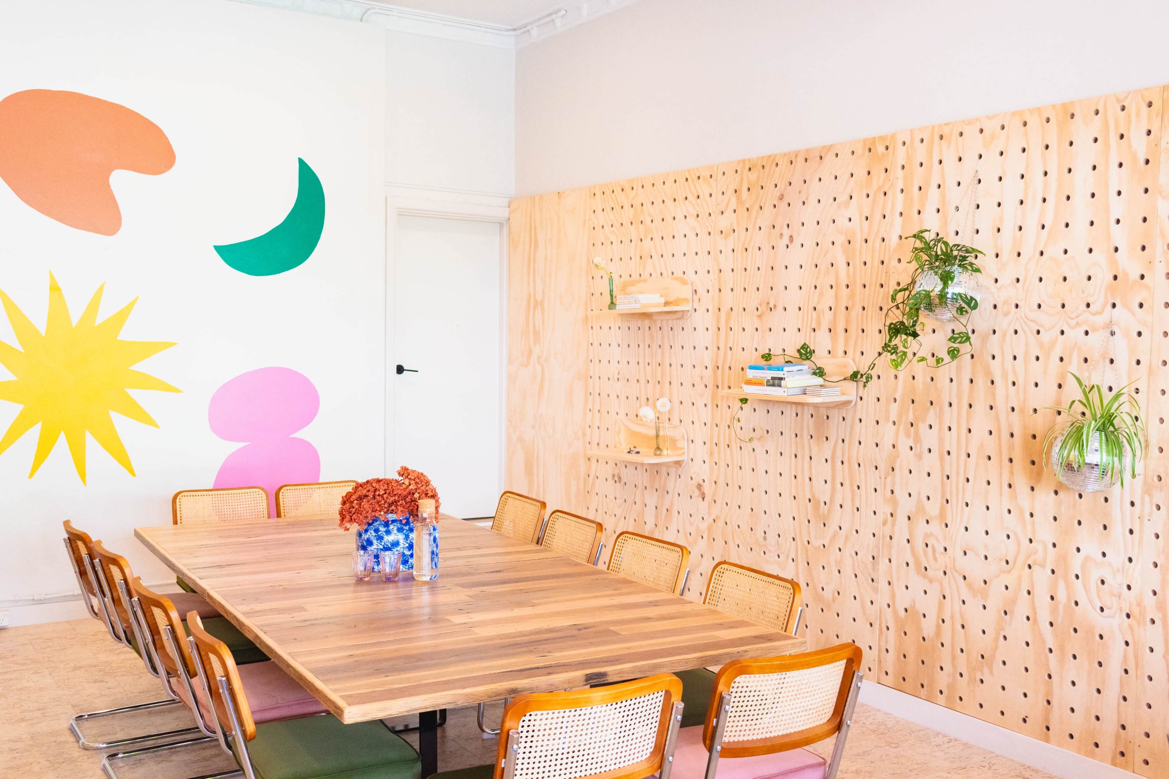 A meeting room features a large wooden table surrounded by colorful chairs, with a wall displaying geometric shapes in bright colors and functional wooden shelving.
