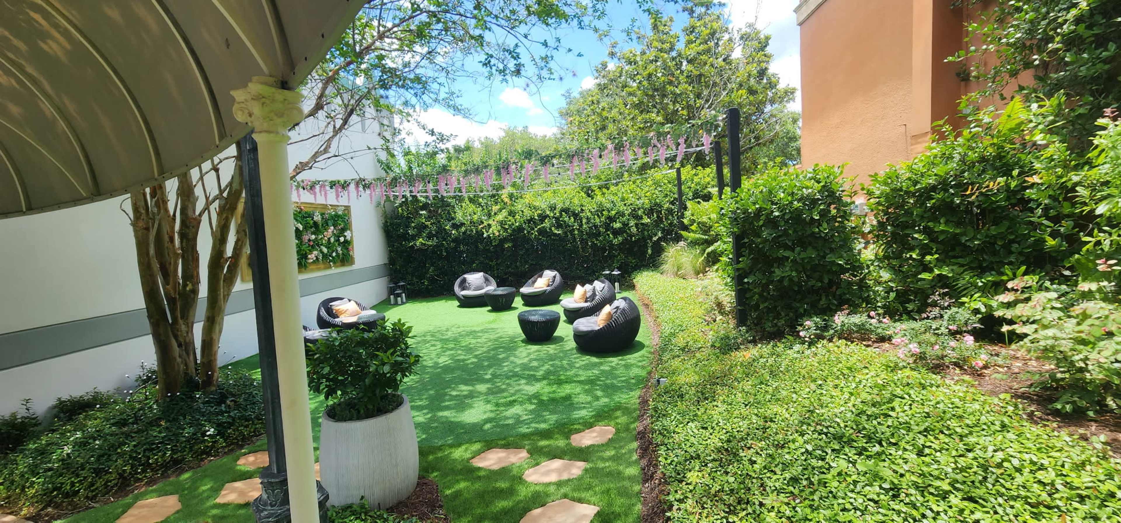 The image shows a landscaped outdoor area with green grass, stone pathways, and black seating surrounded by hedges and trees.