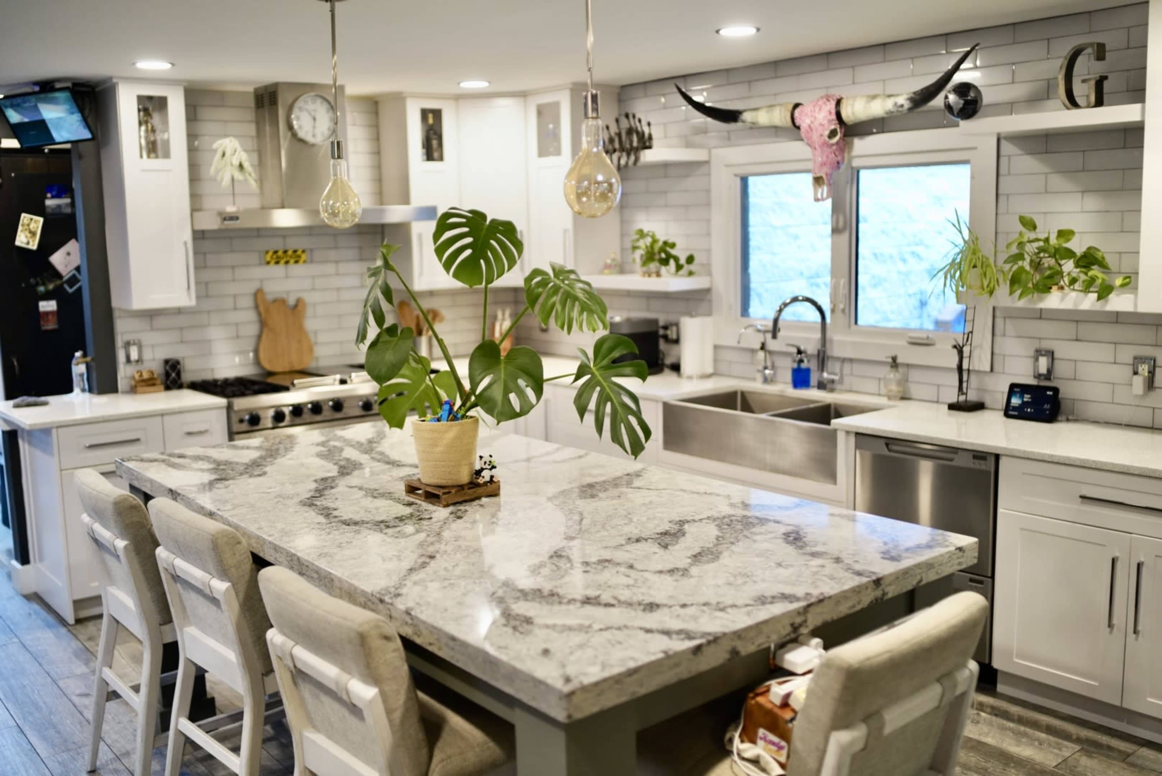 The image shows a modern kitchen featuring a large marble island with seating, stainless steel appliances, and a window overlooking greenery outside.