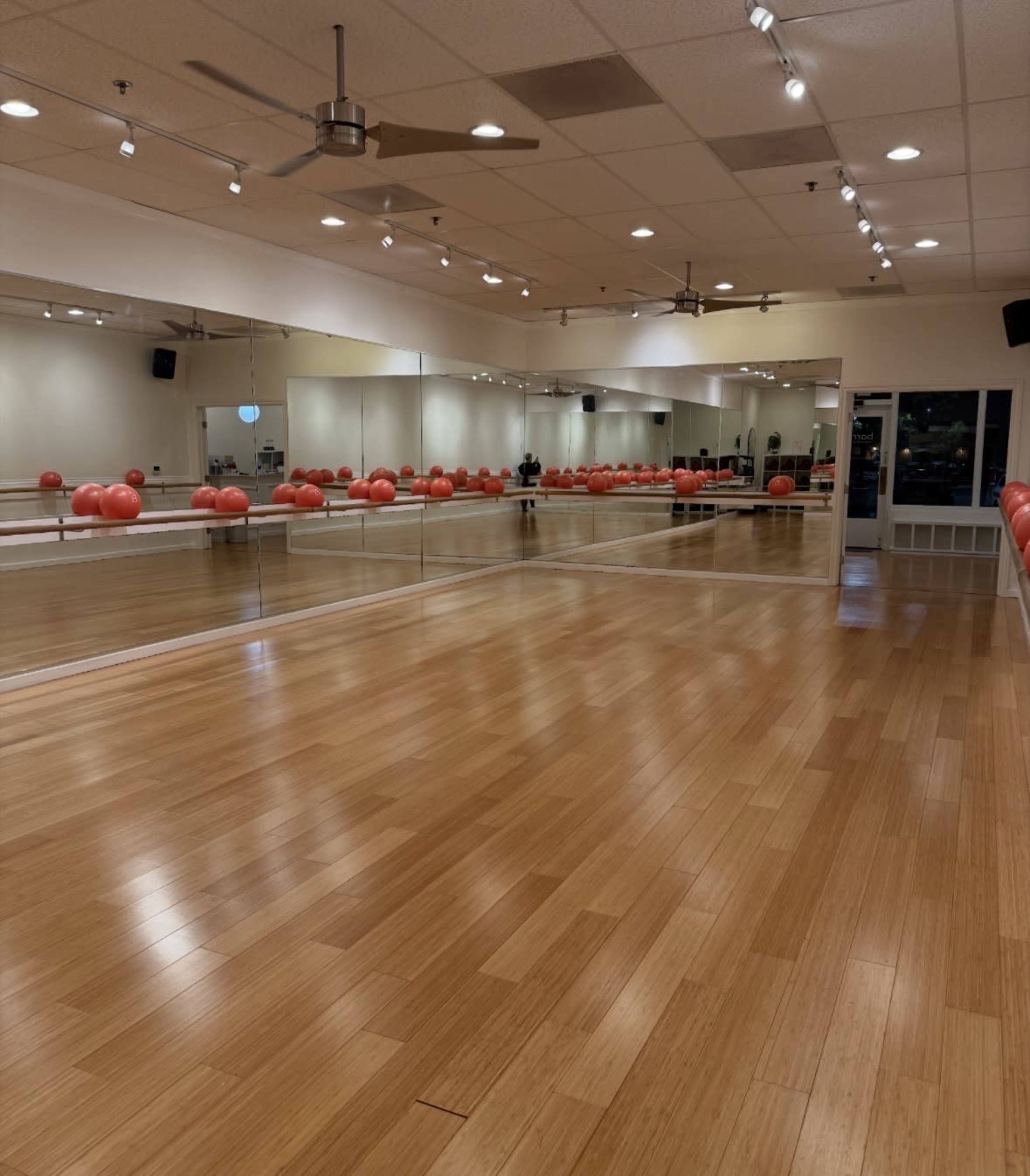 The image shows a fitness studio with a wooden floor, large mirrors, and red exercise balls arranged along the wall.