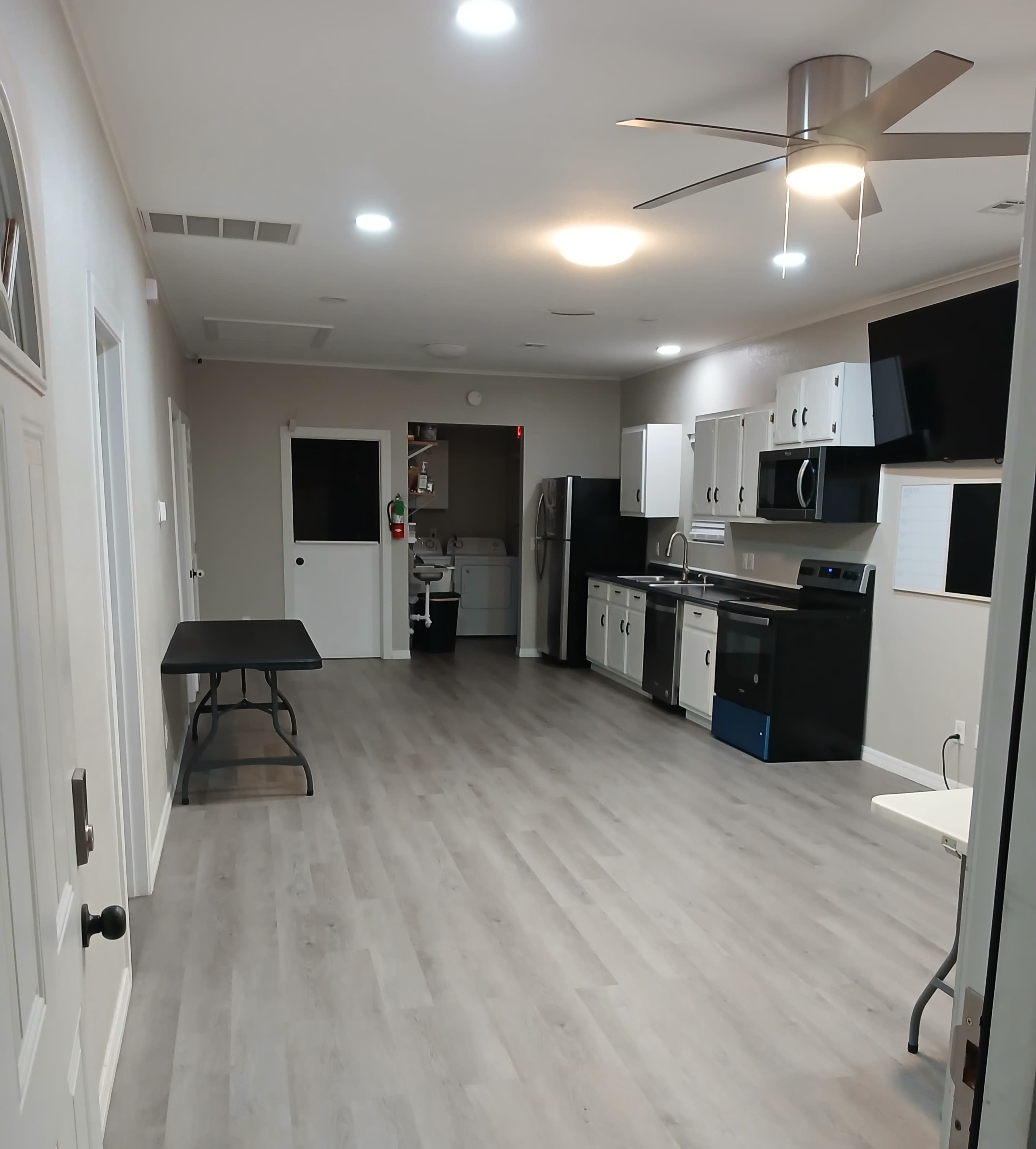 The image shows a modern kitchen with light-colored cabinets, stainless steel appliances, and an open layout featuring a table in the foreground.