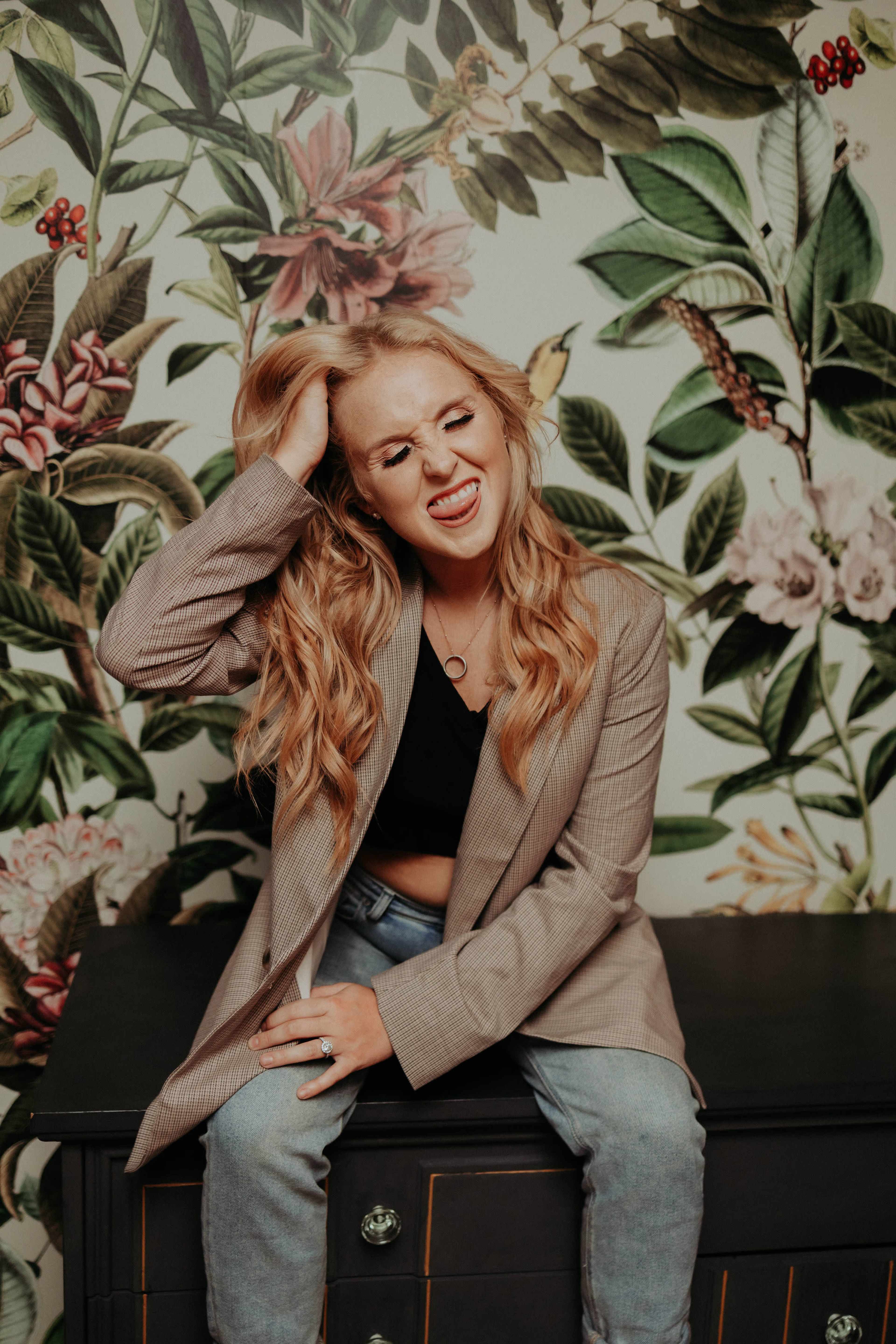 A woman with long hair sits on a dark chest of drawers against a floral wallpaper, pulling her hair and making a playful expression.