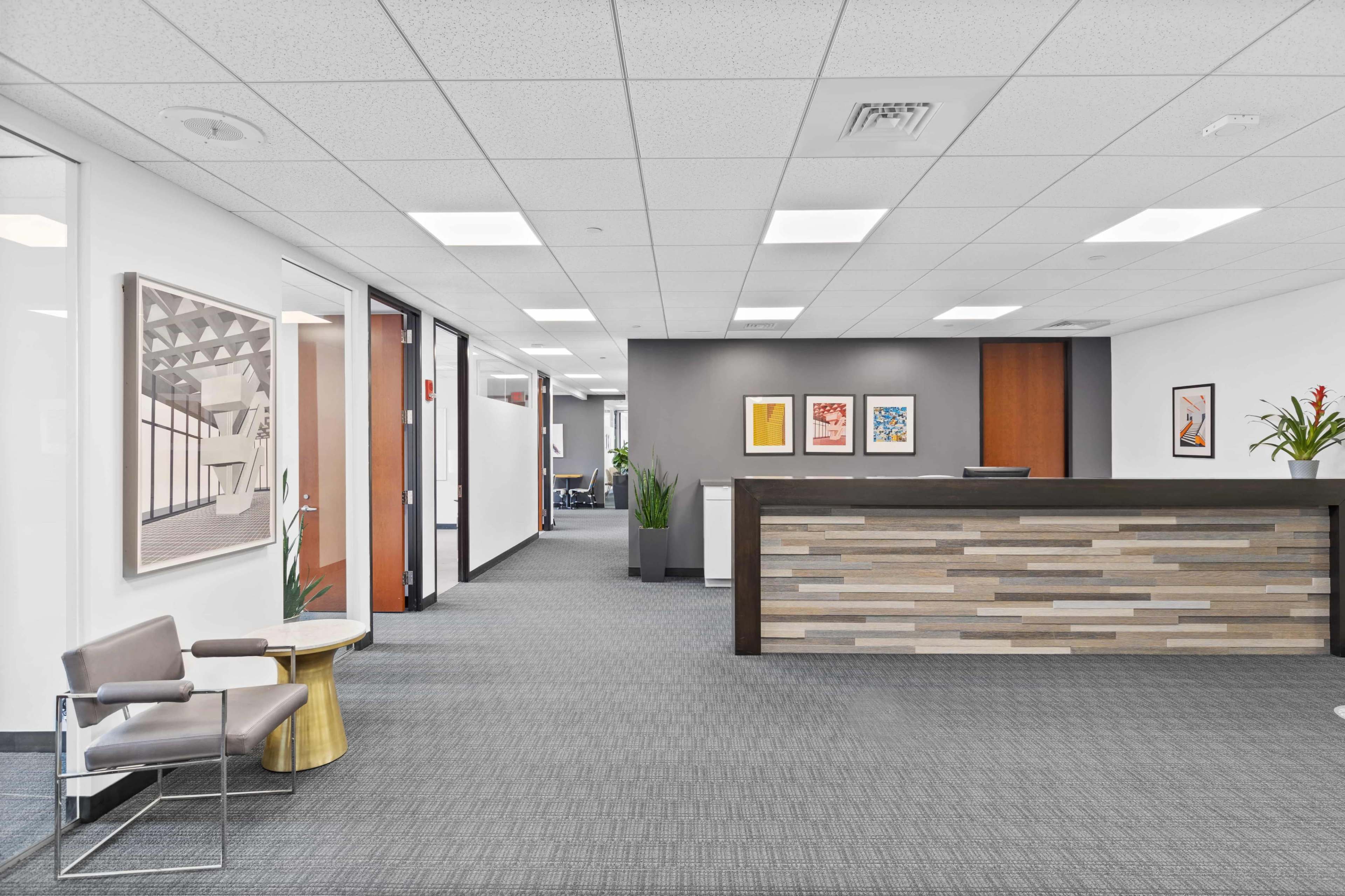 The image shows a modern office interior with a reception area featuring a wooden front desk, artwork on the walls, and a seating arrangement that includes a chair and a small table.