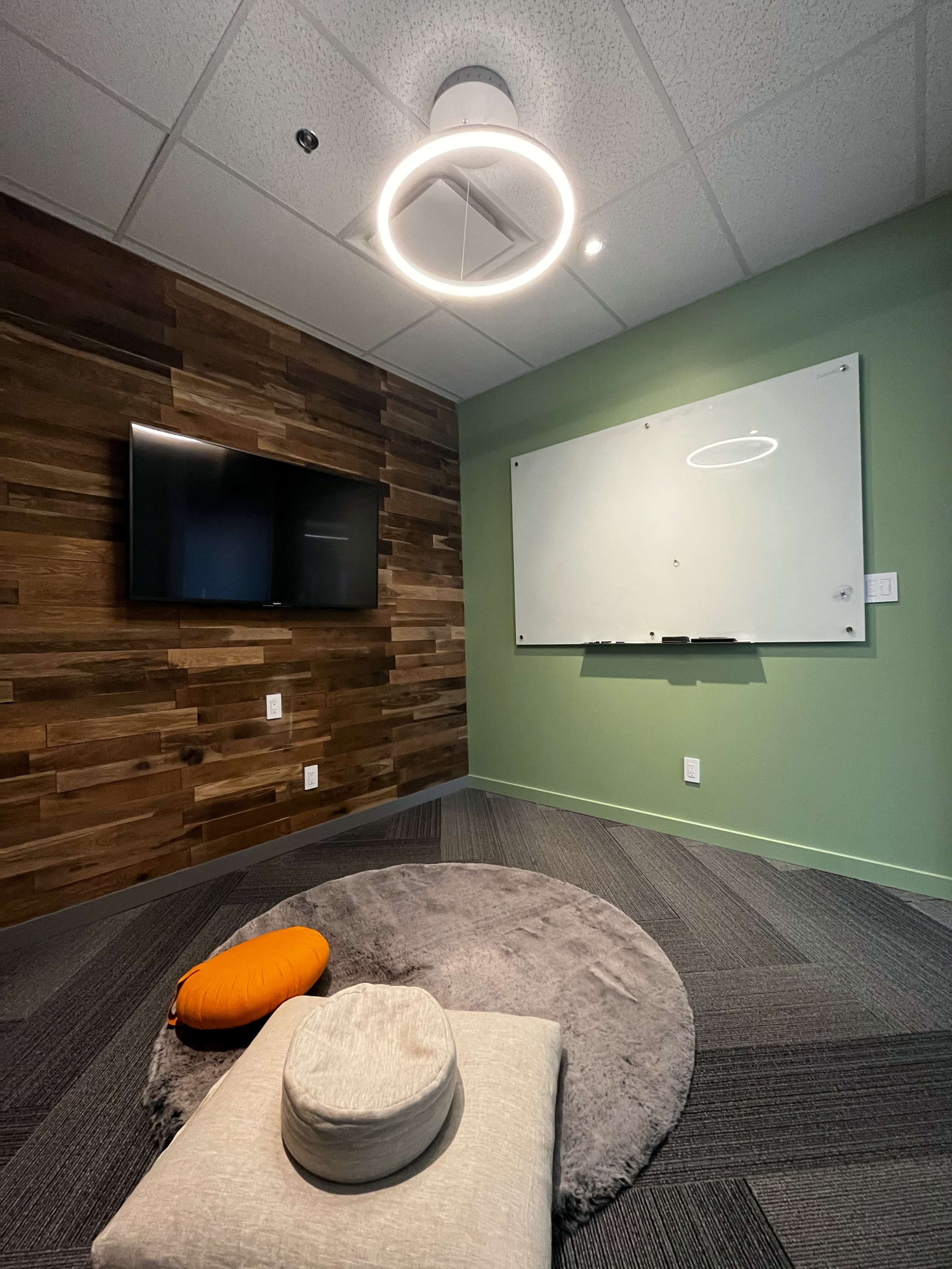 A small meeting room with a wooden wall, a TV mounted on the left, a whiteboard on the right, and floor cushions on a circular rug.