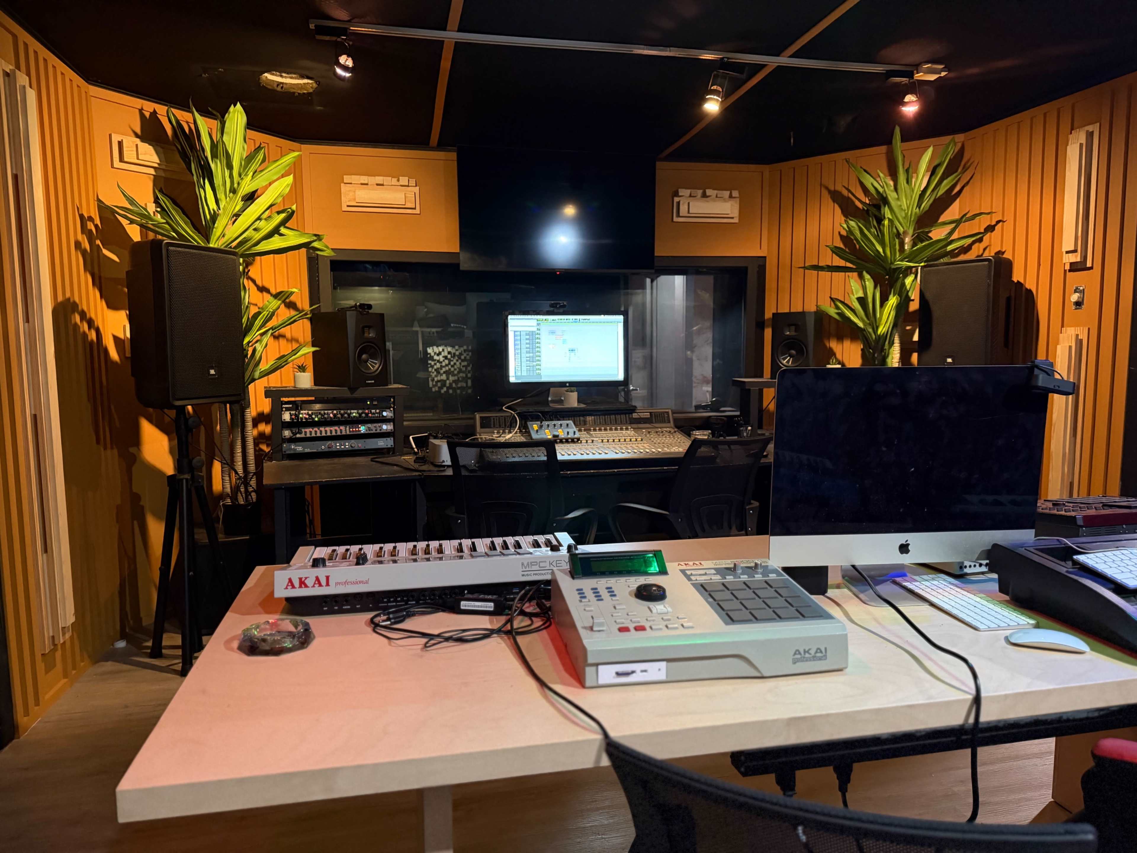 The image shows a music production studio featuring a large mixing console, computer monitor, and audio equipment, surrounded by potted plants and wooden paneling.