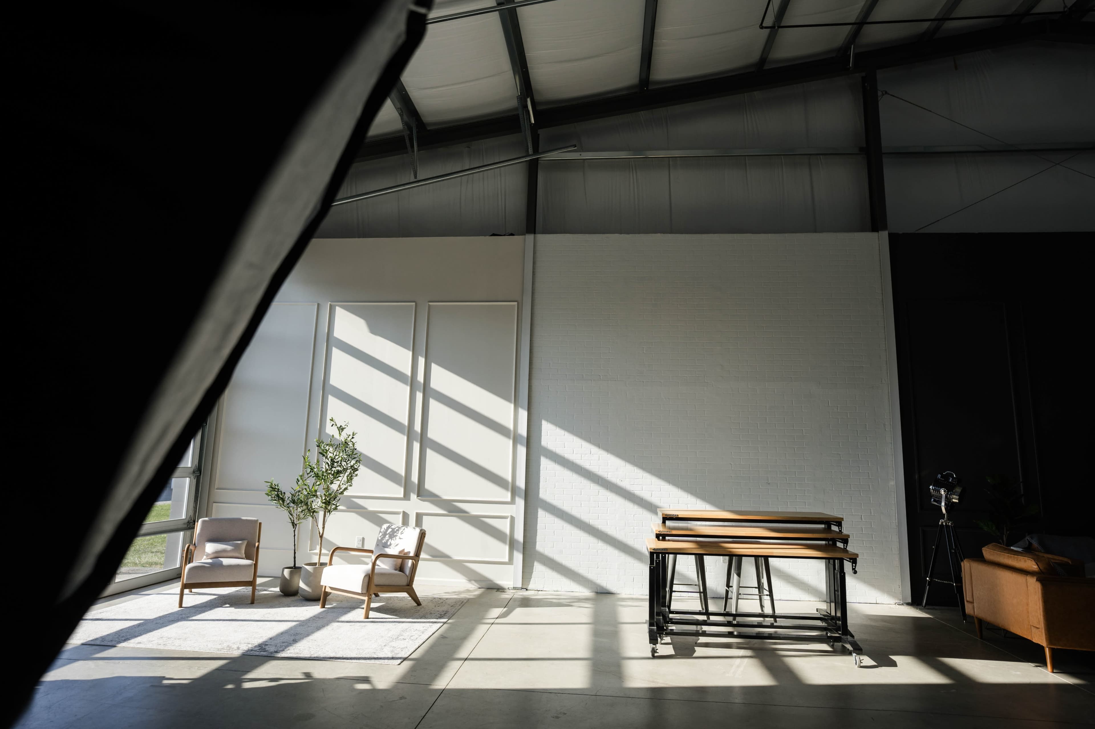 A spacious interior with two chairs, a table on a metal frame, and sunlight casting shadows from large windows onto a predominantly white wall.