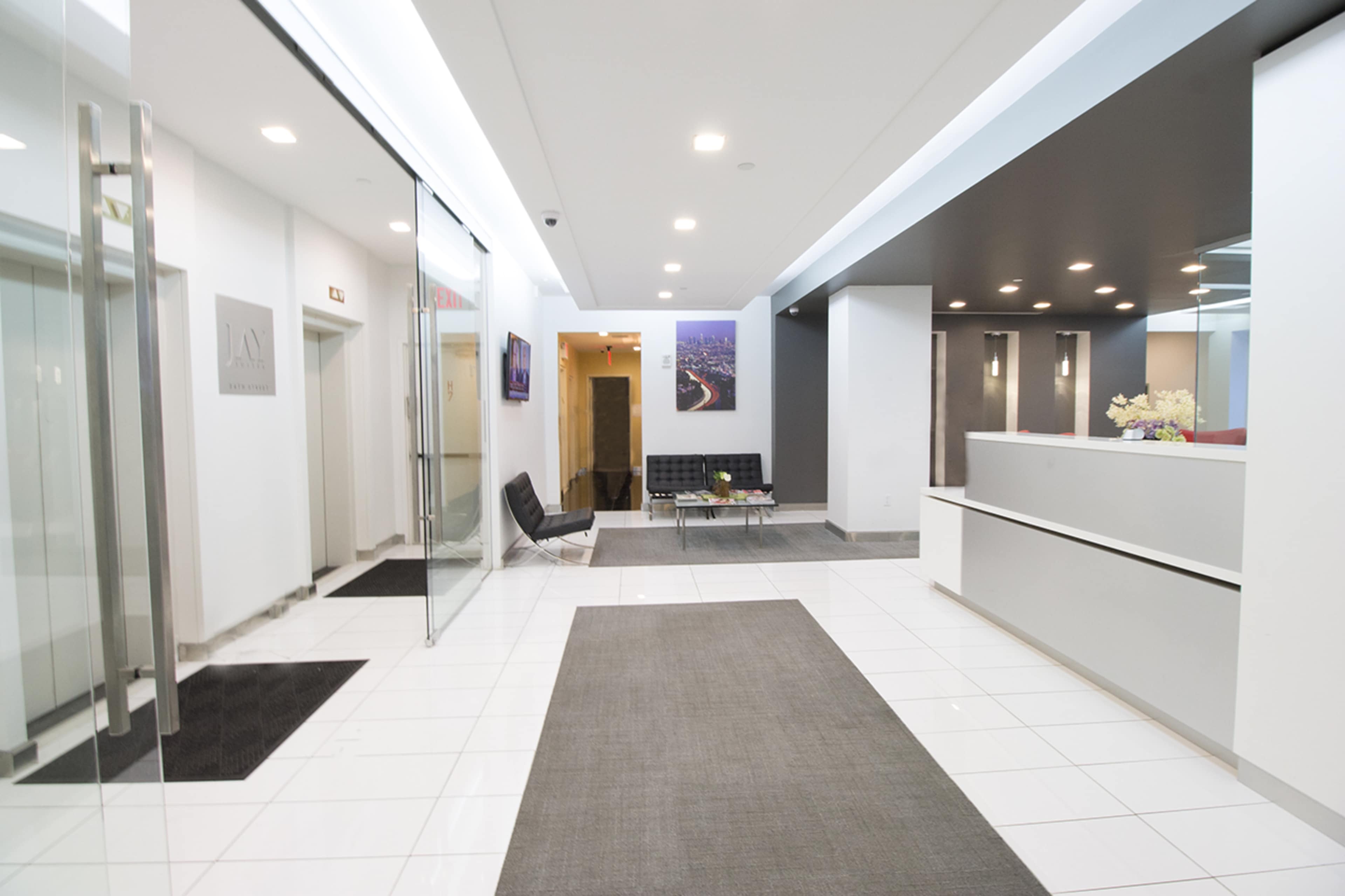 A modern, well-lit reception area with glass elevators, a seating area, and a spacious floor design featuring white tiles and carpet.